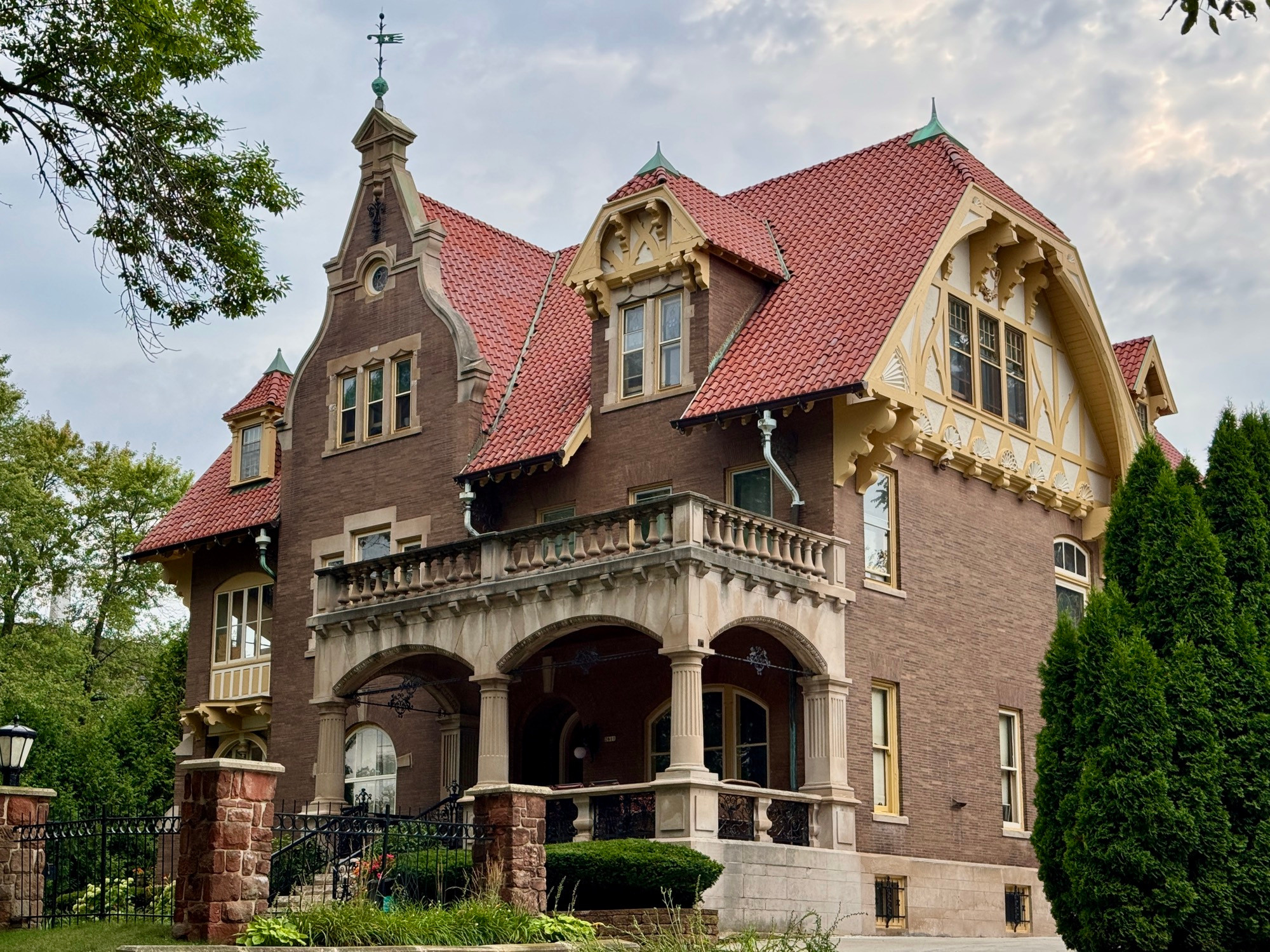 A giant Germanic megabungalow with red tile roof, crazily ornate gables and dormers, a columned porch, an iron fence, a weathervane, and seashell details along side gable