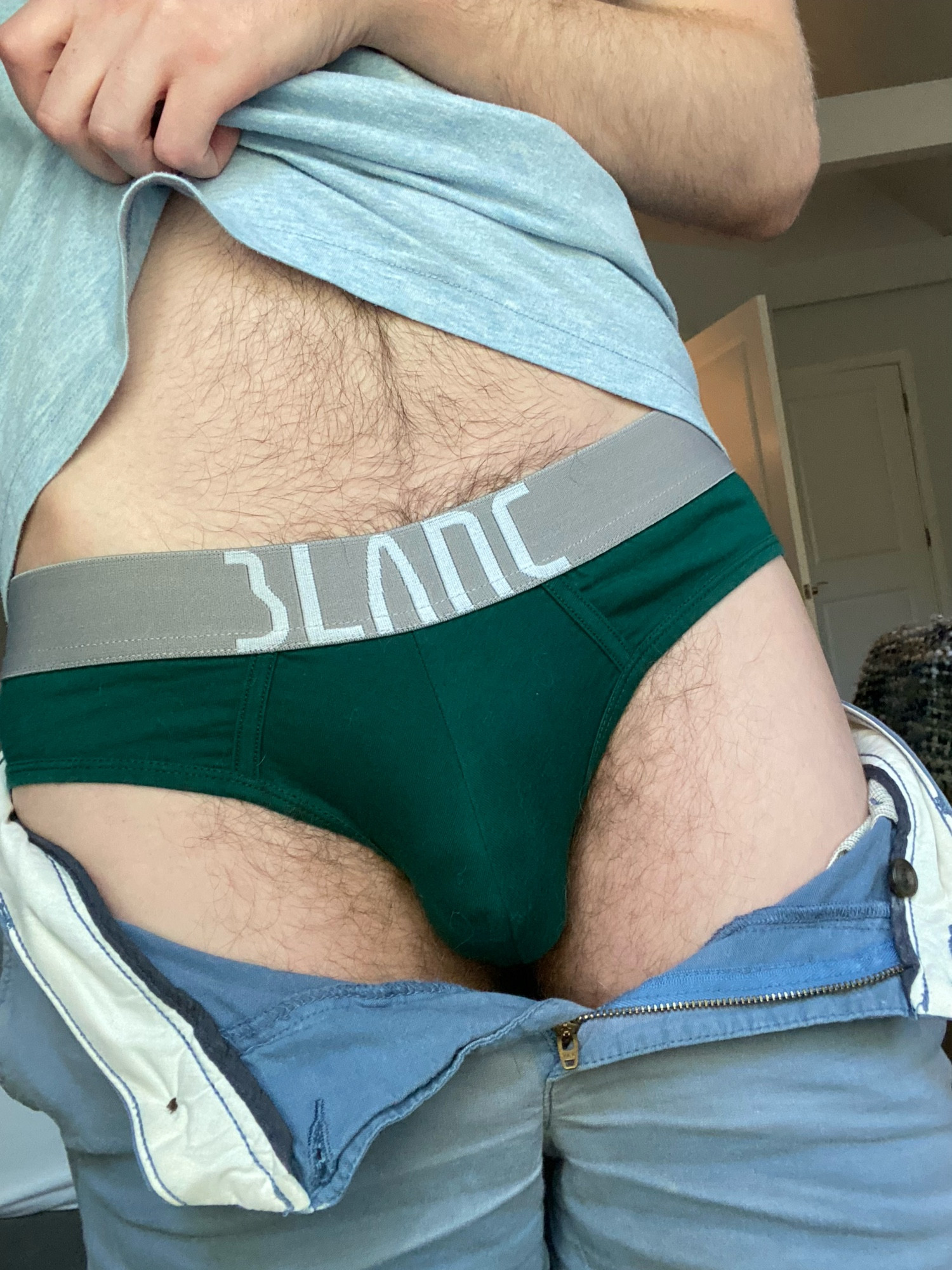 Photo of Happy from navel to thigh. He is pulling up his shirt to expose his happy trail reaching down to forest green briefs as his shorts are unbuttoned showing the slightest hint of his cock head.