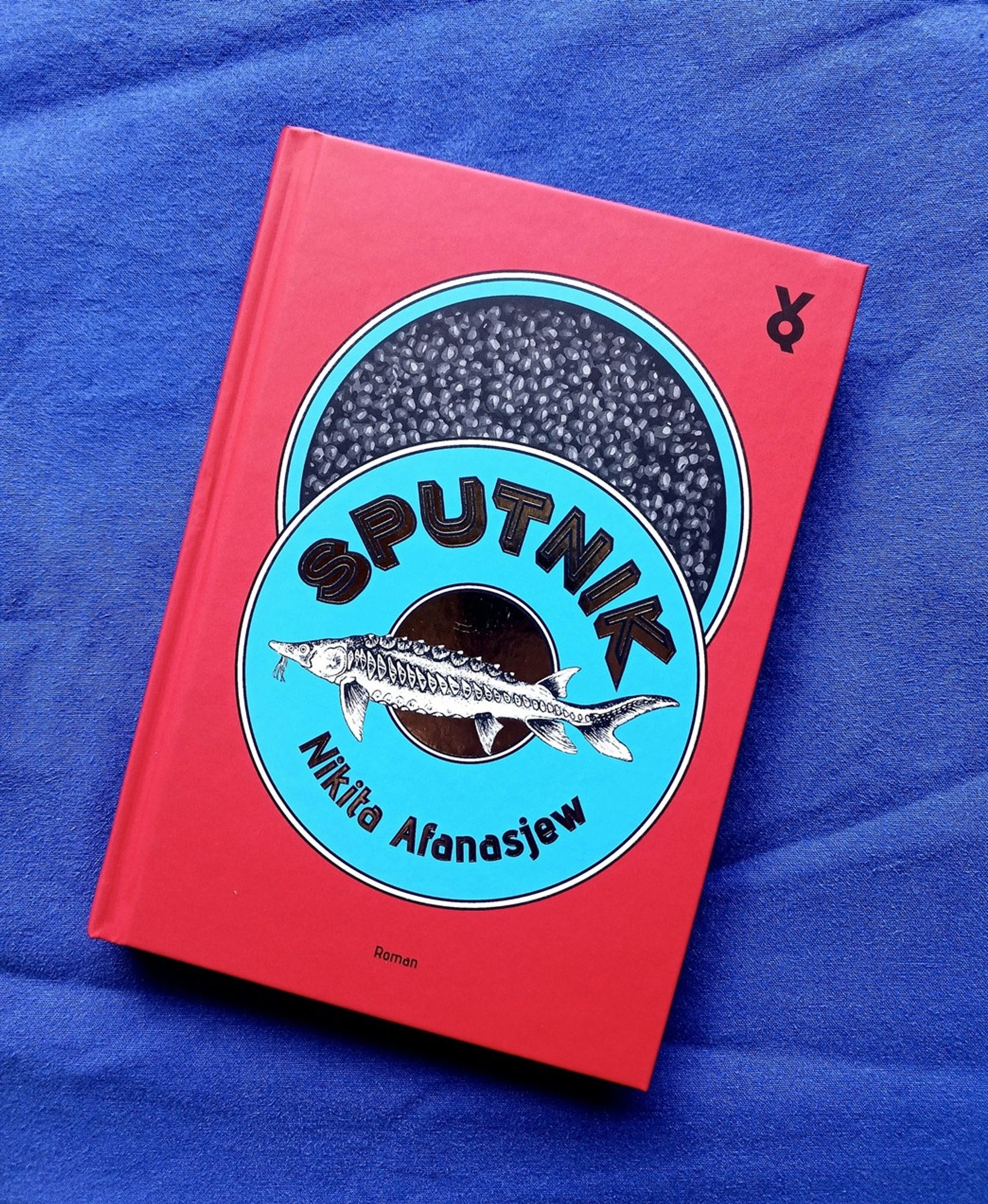 Foto des Buches, auf dessen rotem Cover die Zeichnung einer geöffneten blauen Kaviardose zu sehen ist, auf der Autorname und Titel rund um einen Fisch als Aufschrift zu lesen sind.