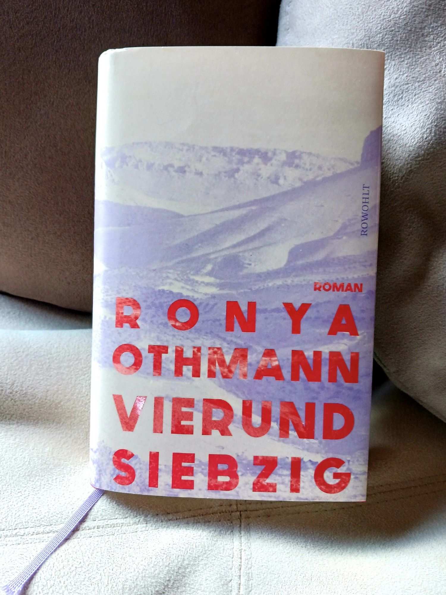 Foto des Buches, auf dessen sandfarbenem Cover eine hellviolette karge Wüsten- und Gebirgslandschaft zu sehen ist. Der Name der Autorin und der Titel des Buches stehen am unteren Rand in großen, rot glänzenden Buchstaben. Das Buch steht auf einem hellen sandfarbenen Sofa gegen zwei Kissen in unterschiedlichen Brauntönen gelehnt.