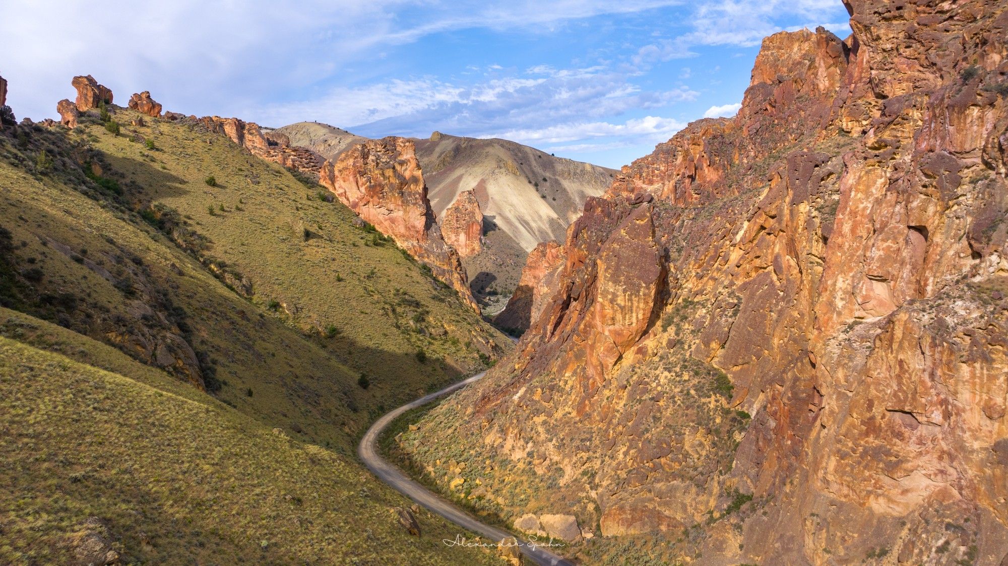 A winding dirt road cuts through a gulch with green vegetation and towering golden rocks on both sides.