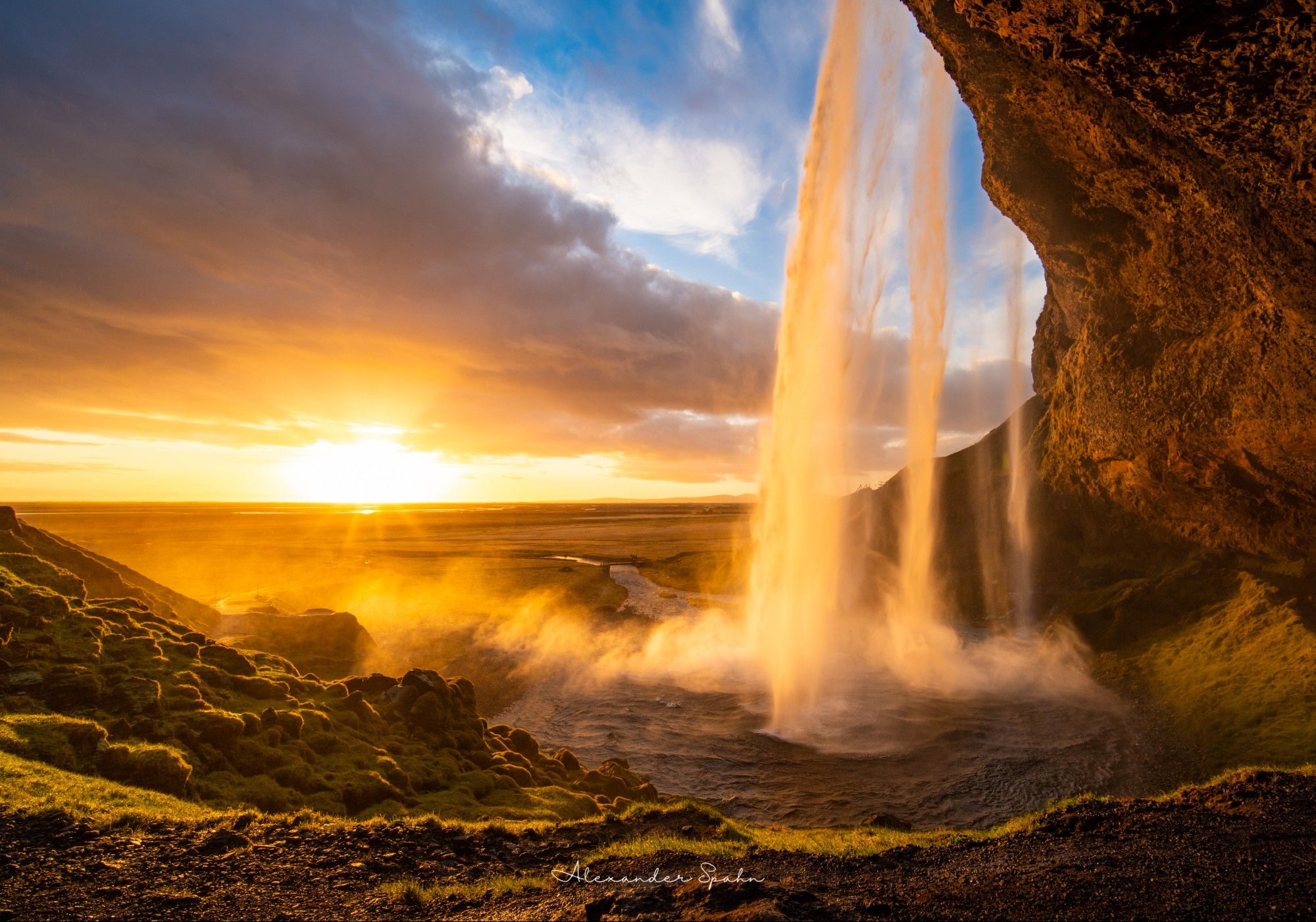 A waterfall is illuminated from behind by the Sun, which is setting on the distant horizon below a layer of clouds.