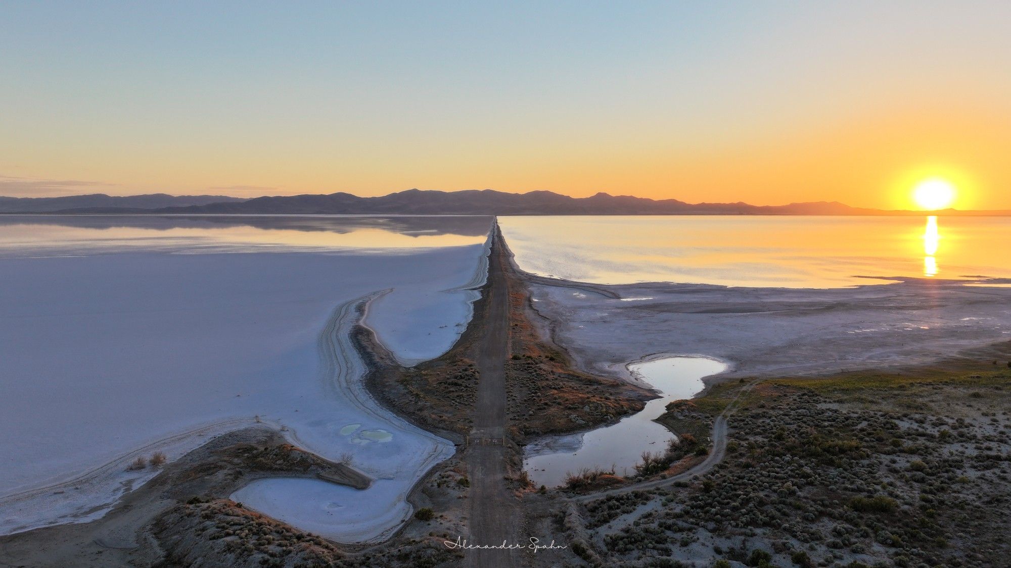 A long road extends out between two large bodies of water. The Sun sets behind a distant mountain range.