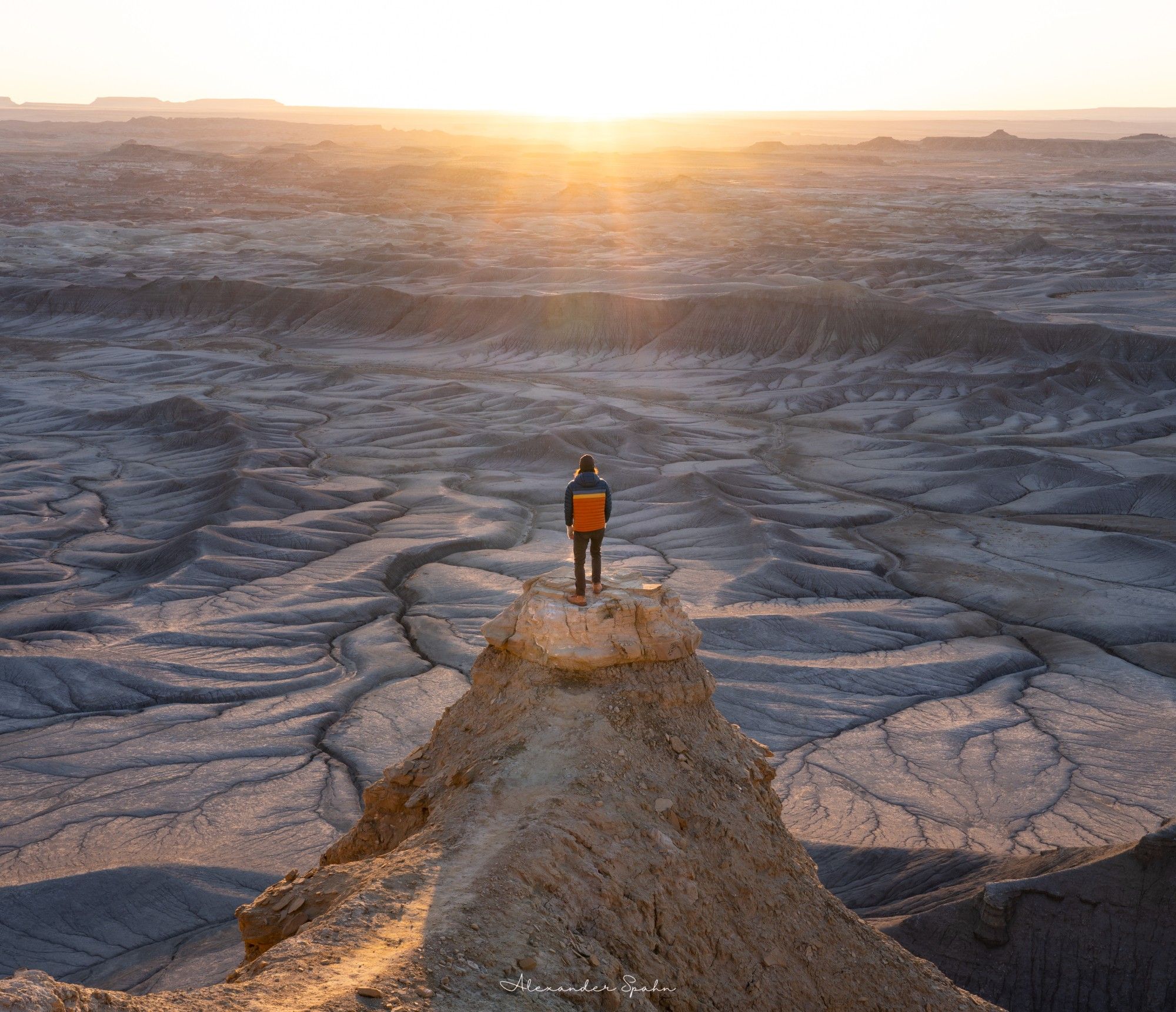 A selfie (from a remote camera) of me standing on the edge of a very narrow, ~1,5000 ft cliff overlooking a Moon-like desert landscape as the Sun rises in the distance.