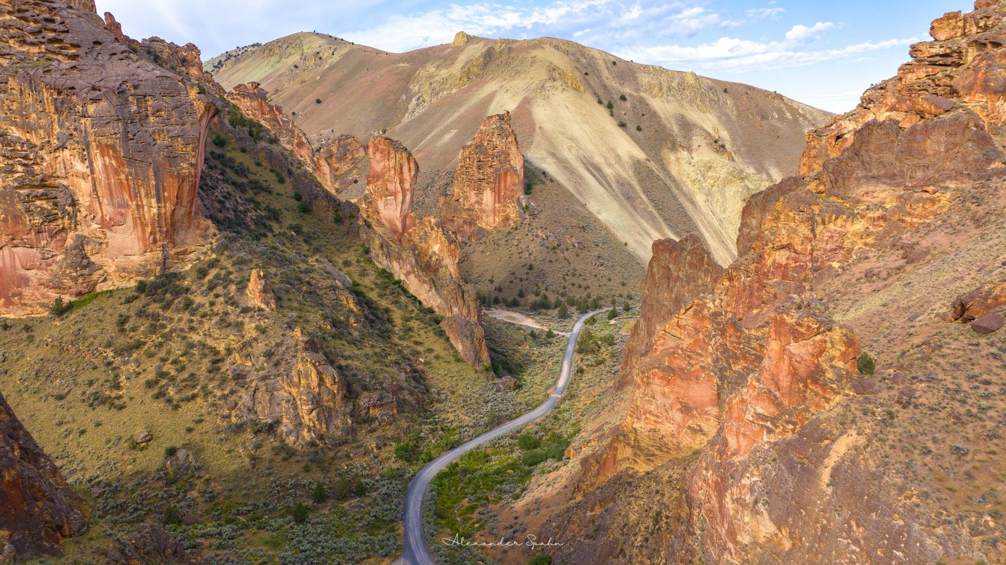 A winding dirt road cuts through a gulch with green vegetation and towering golden rocks on both sides.