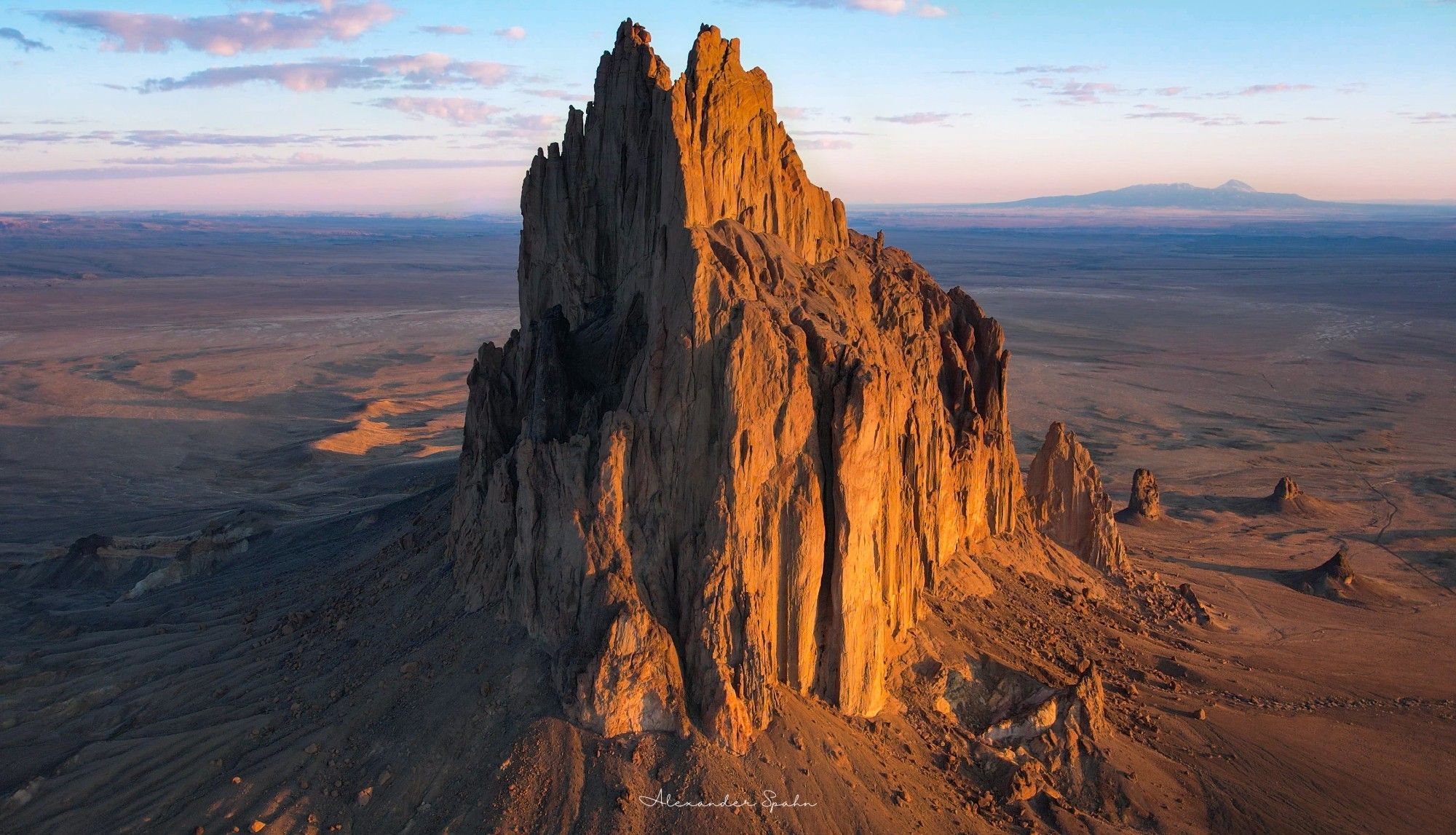 A sunlit Shiprock sticks out of the ground at sunrise, with pastel color skies and clouds above and a very distant mountain in the background.