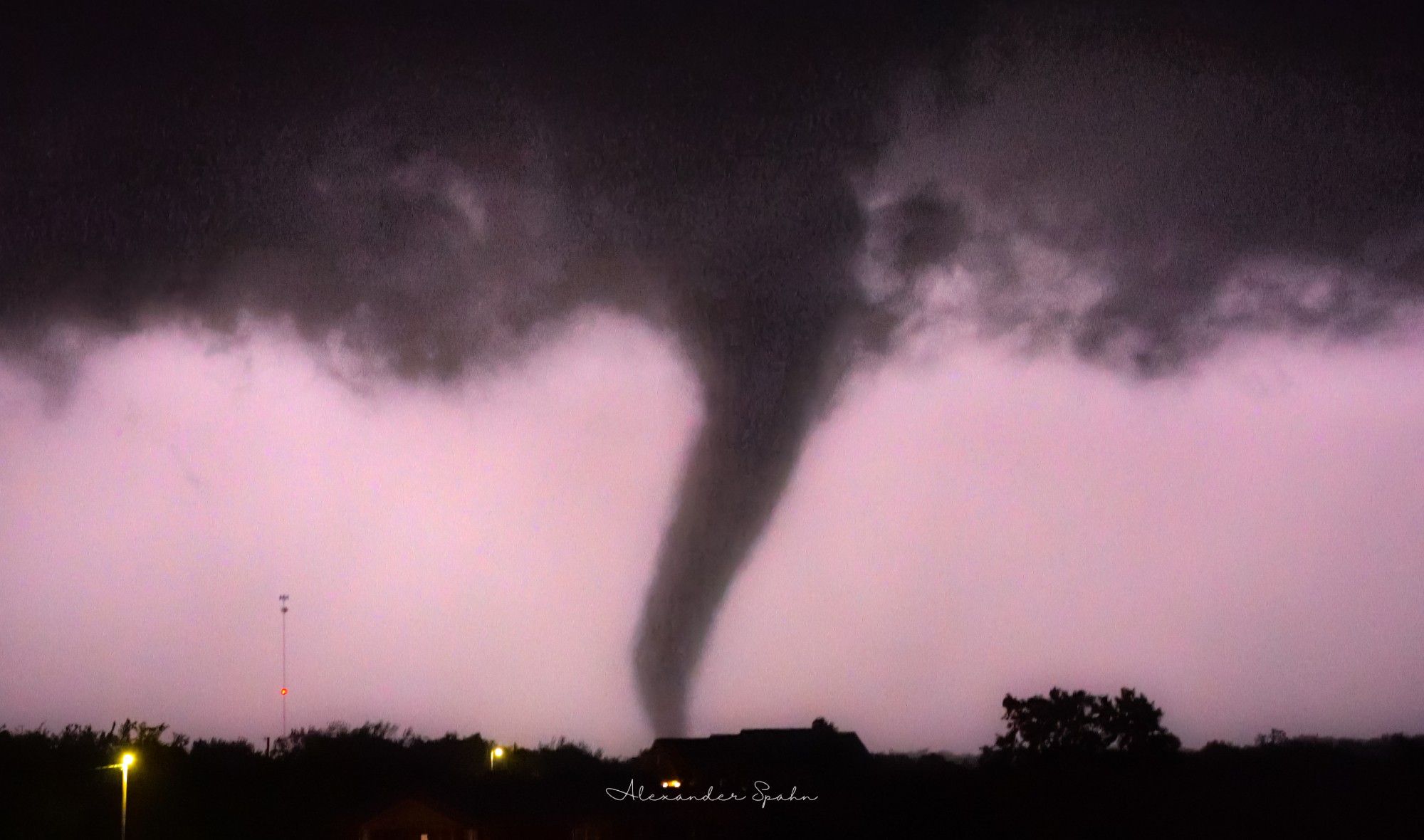 A tornado after dark is center frame with the background sky illuminated a slight purple by lightning. Trees and houses are in the foreground.