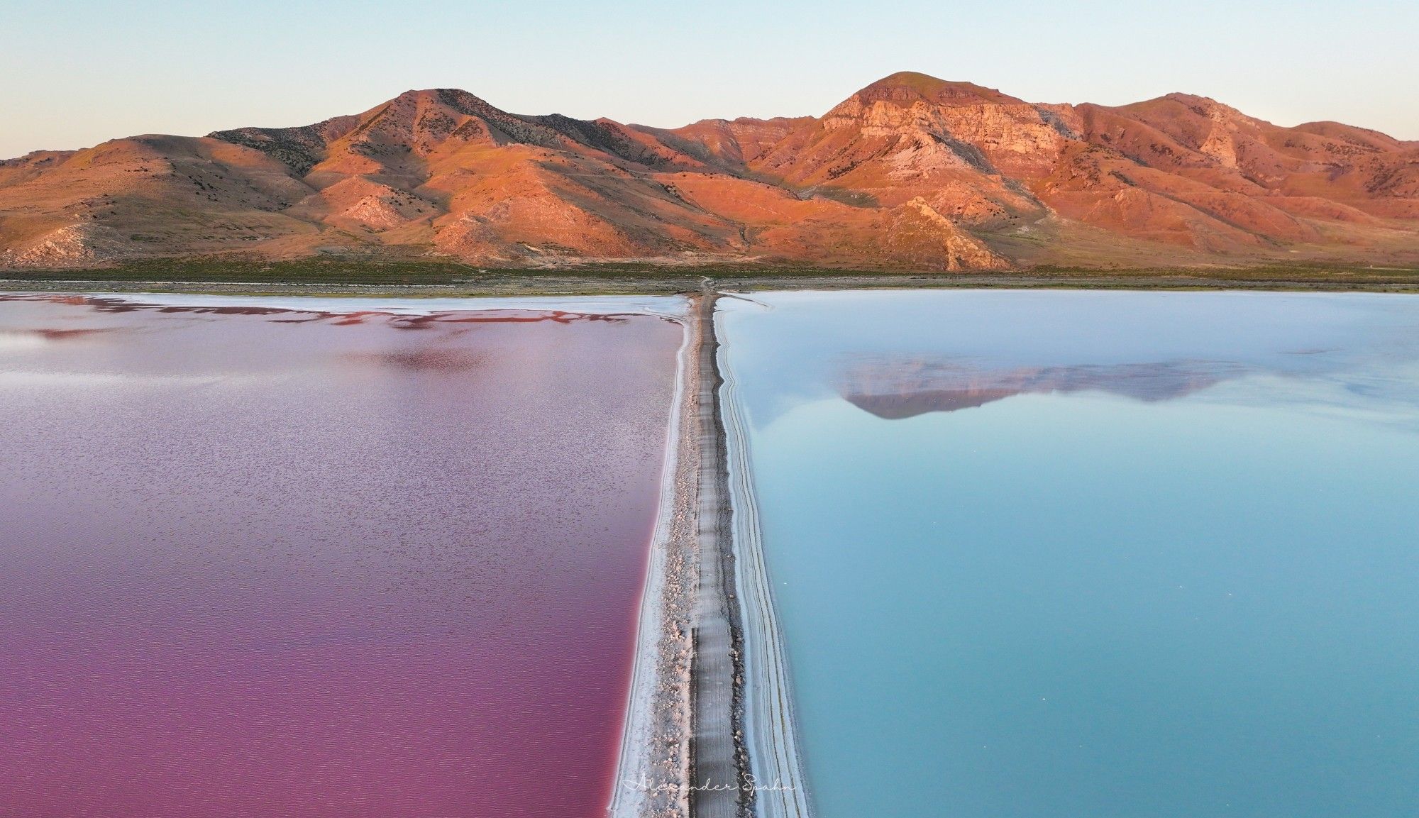 A lone white road separates blue waters from pink waters, with sunlit mountains at the end of the road.