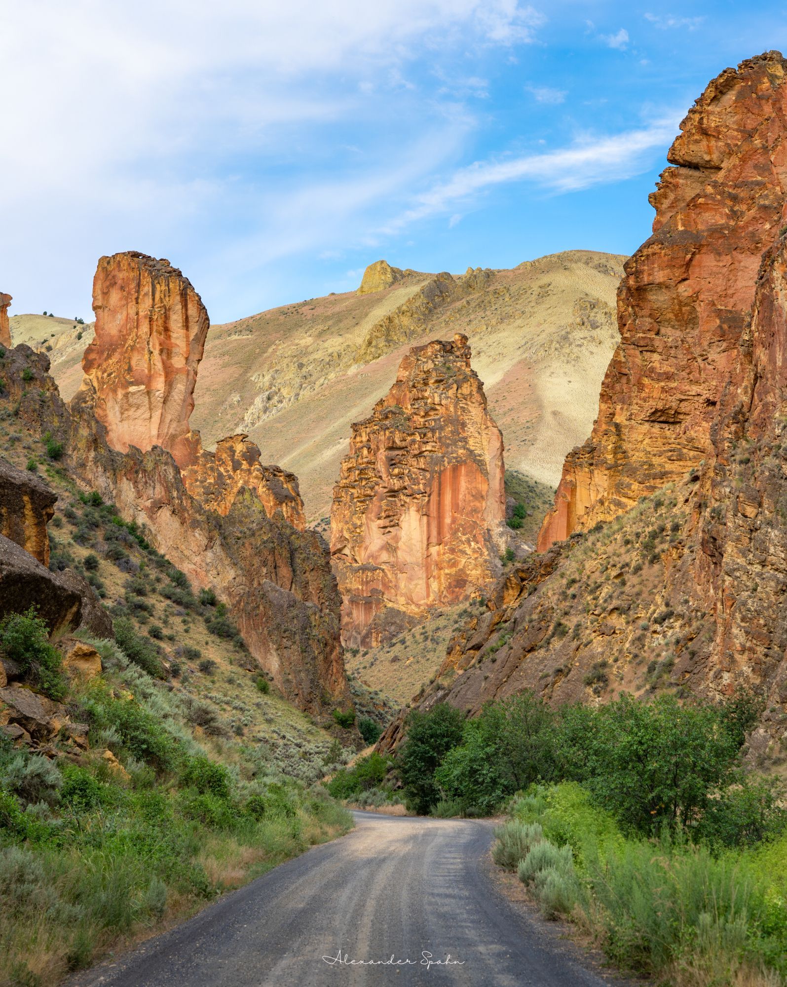 A gravel road extends into a winding gulch, bracketed by steep slopes and towering orange/golden rocks.