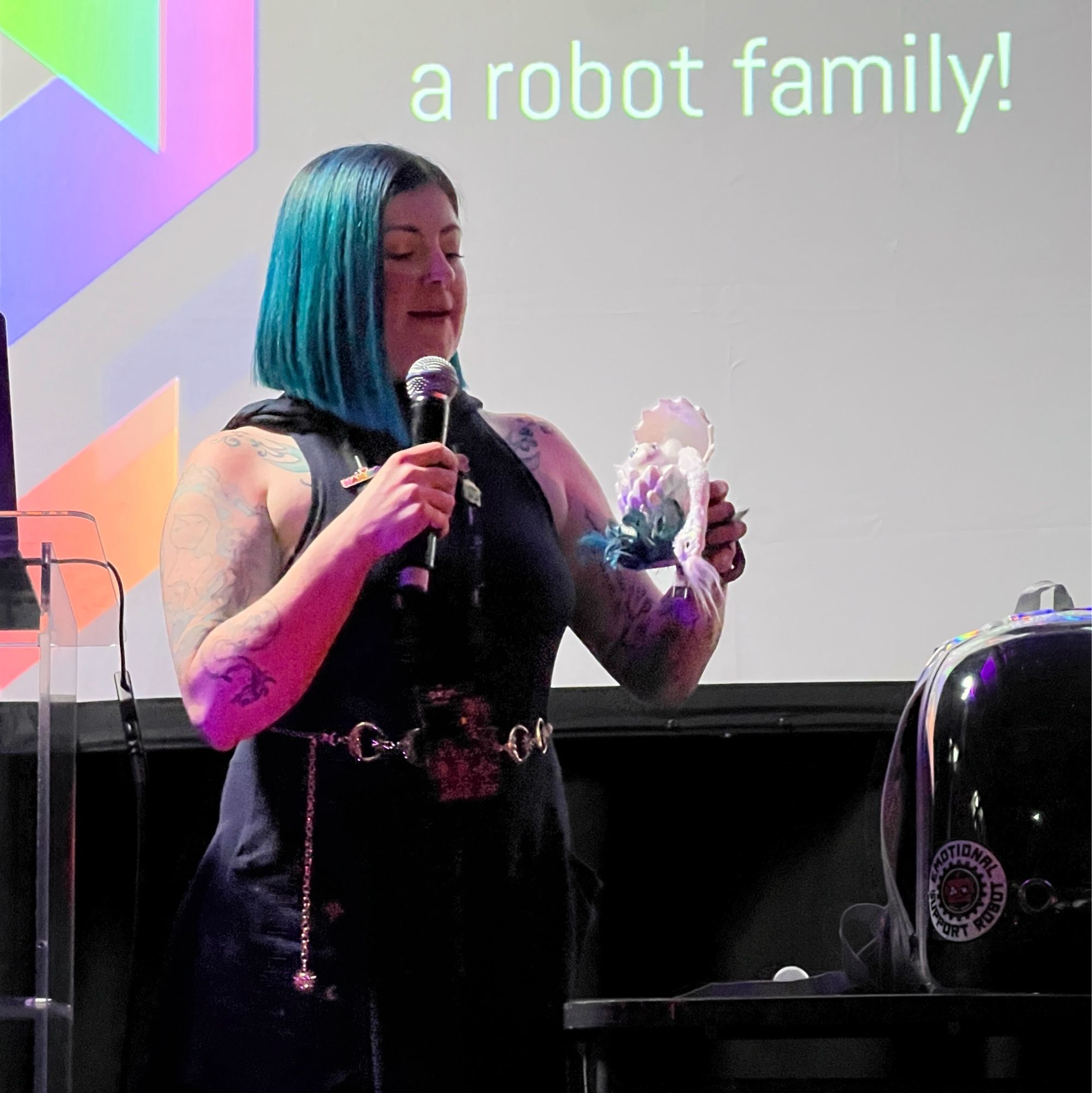 Angela shows a little dragon/fairy egg bot, on stage at Supercon '23. Its display-case backpack sits on a table.