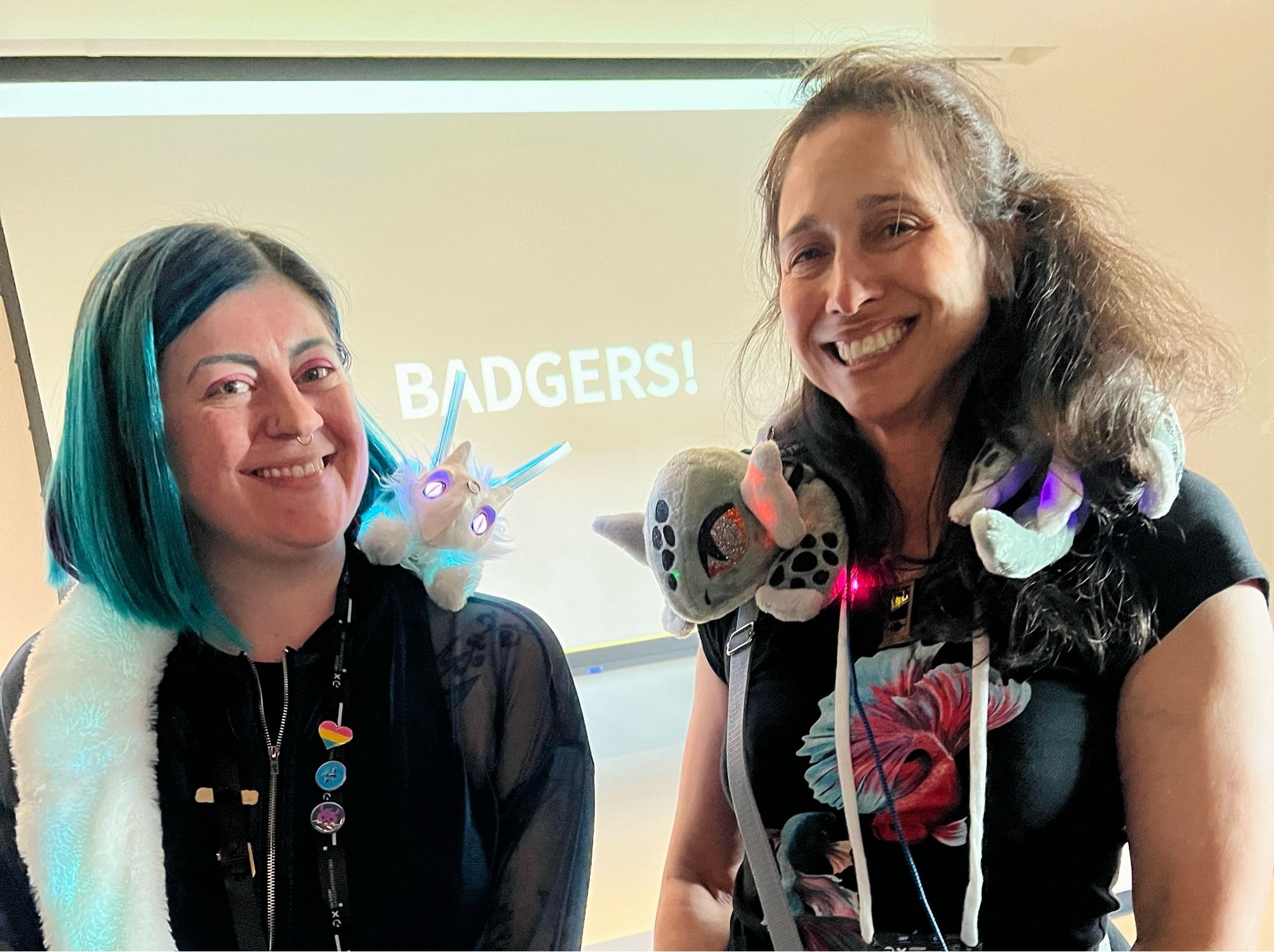 Angela Sheehan (GellaCraft, left) with her little furry dragon-fairy robot, Nova; and Debra Ansell (GeekMomProjects, right) with her grey plushy light-up bot. Both bots are draped across their makers' shoulders.