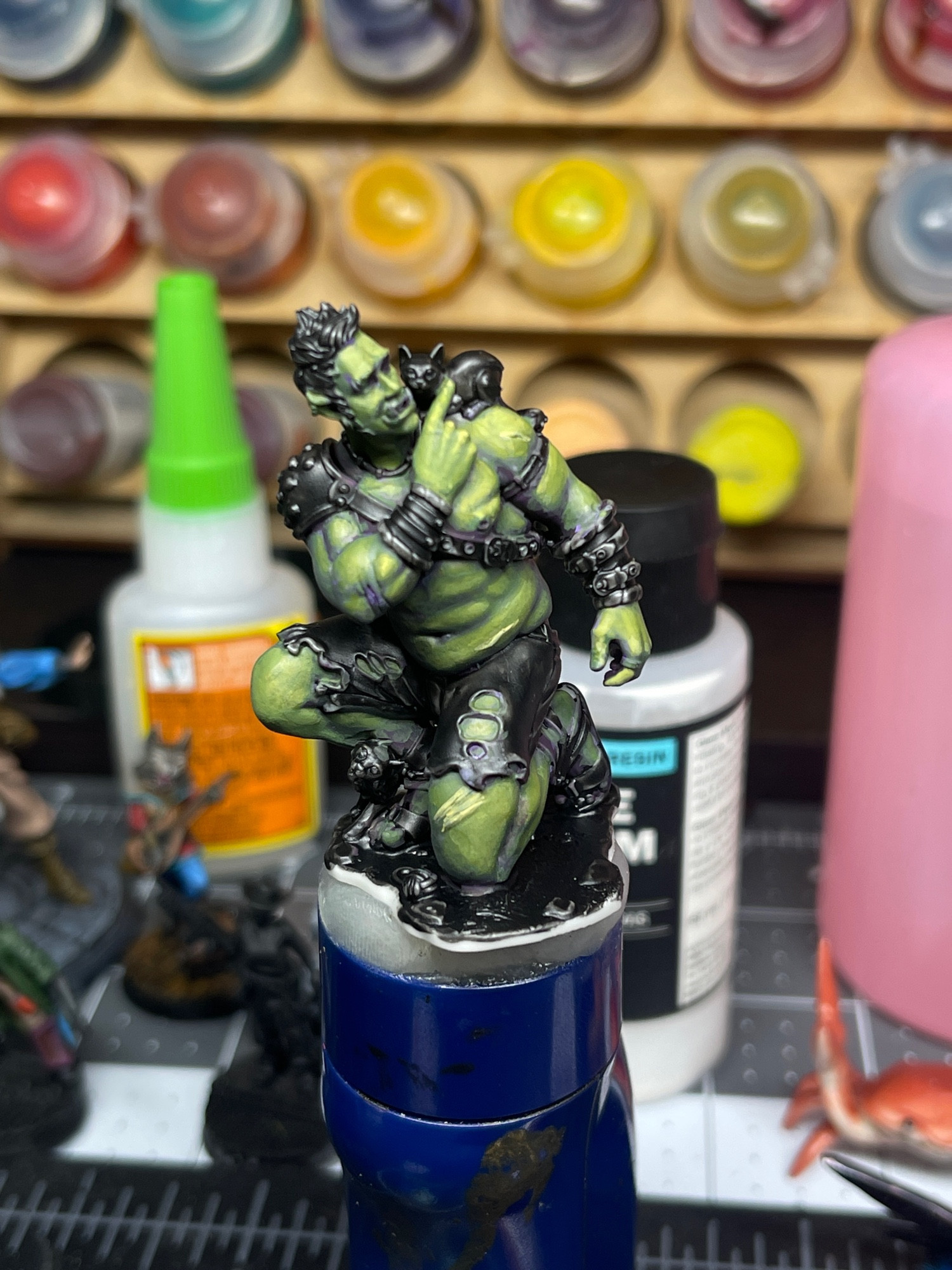 A miniature figure of a strong, but soft looking male half Orc. Wearing shorts and no shirt, he is on one knee with his right hand crossing his body to the top of his left shoulder to pet a small cat perched there. Under his right knee, another cat rubs against him. Only his skin is currently painted with green and yellow highlights, and purple shadows where his muscles are most prominent.