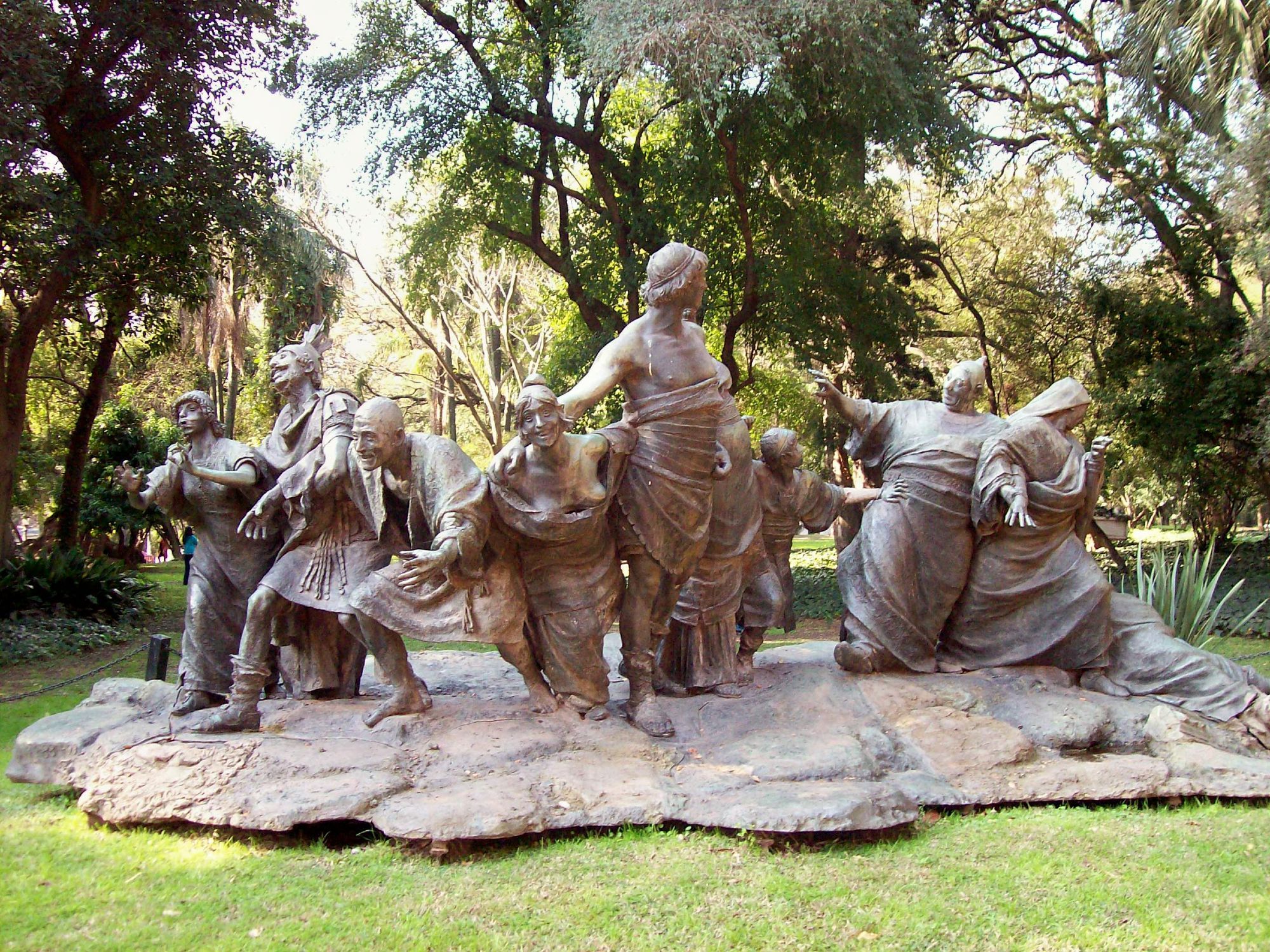 9 bronze figures, mixed male and female, in ancient Roman dress stumble joyously together; photograph of 1909 copy of 1899 brown sculpture "Saturnalia" by E. Biondi courtesy R. Fiadone via Wikimedia Commons (CC-BY-SA-2.5)