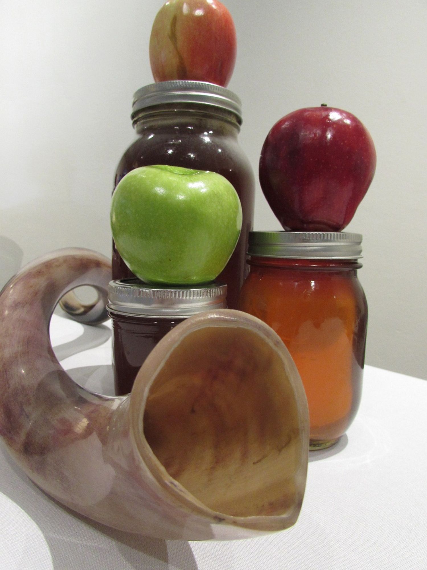 A photographic still life of a shofar, or ram's horn, with three apples and three jars of honey arranged on a white table against a white background, image courtesy cstpdx via Pixabay