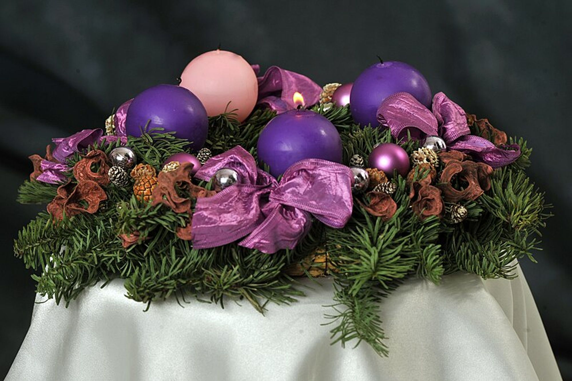 Advent wreath with 3 purple and 1 pink round candles with purple ribbons and red and silver balls among the evergreens sitting on a white table cloth against a dark background; the foremost purple candle is lit, courtesy Philip János (public domain) via Wikimedia Commons