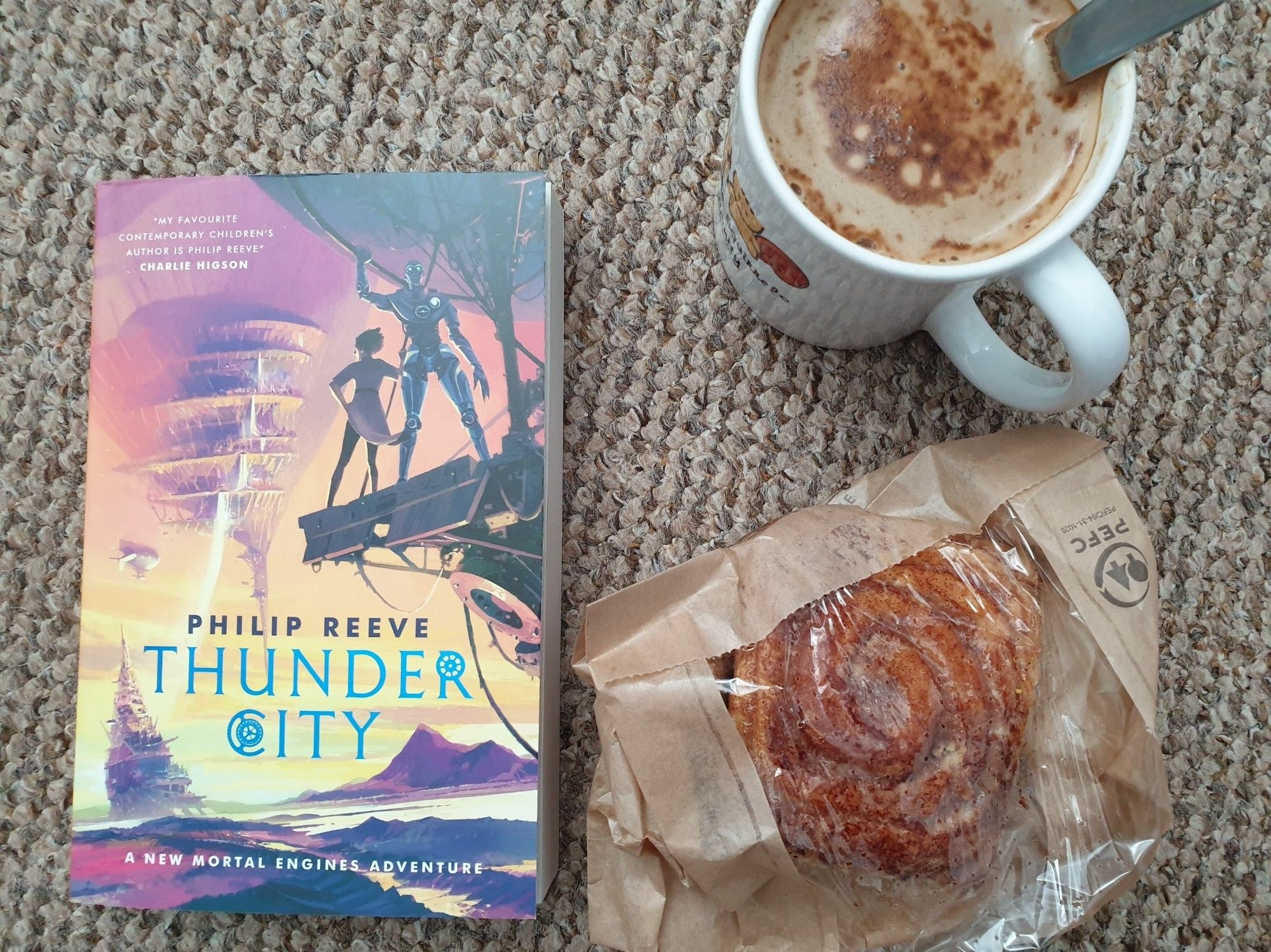 On my carpeted floor, three of my most favourite things. A cinnamon bun, a cup of coffee, and the new Mortal Engines book by Philip Reeve.