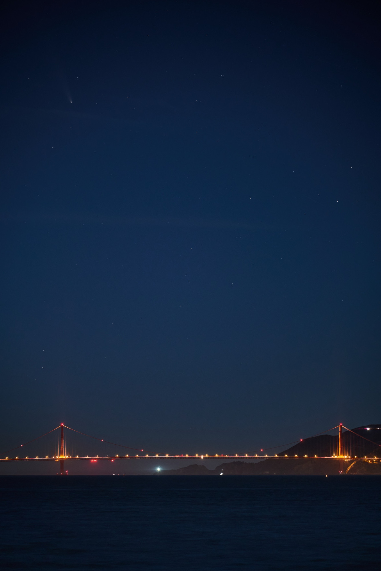 A brightly lit suspension bridge spans across a body of water at night, with stars visible in the sky above. A comet can be seen in the upper left corner, adding to the scenic view.