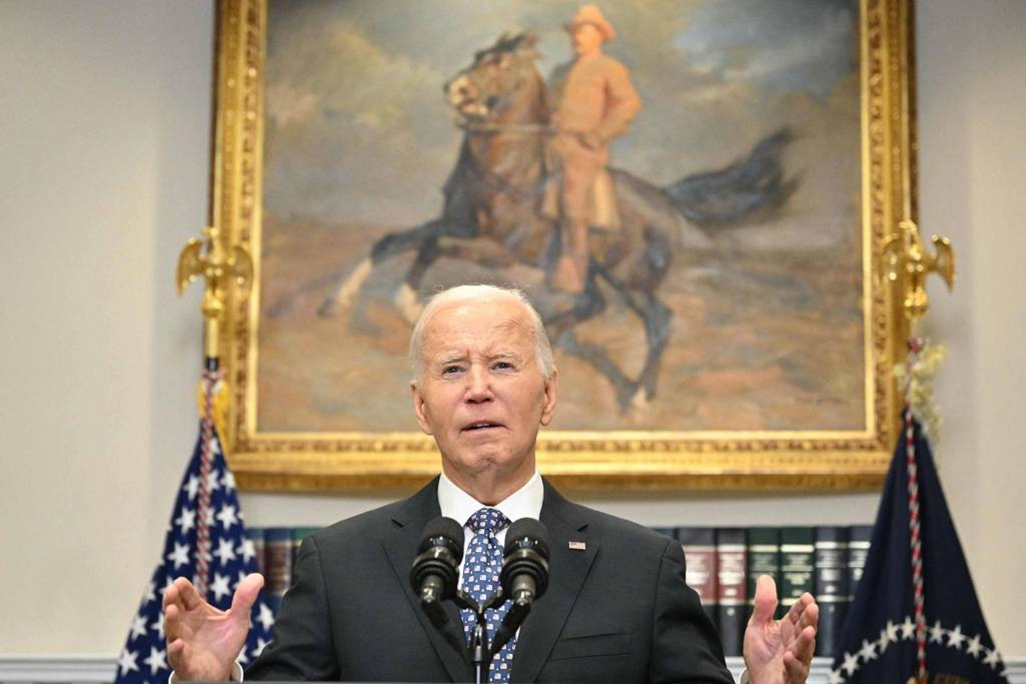 Fotografia colorida é retrato do presidente dos EUA Joe Biden, visto da cintura para cima. Biden é um homem branco, de cabelos brancos, lisos e curtos, e está vestindo terno cinza, camisa branca e uma gravata azul com desenhos brancos. Ele está discursando em frente a um microfone, e está gesticulando com as duas mãos, com as palmas abertas. Atrás dele, há um quadro antigo de um soldado à cavalo.