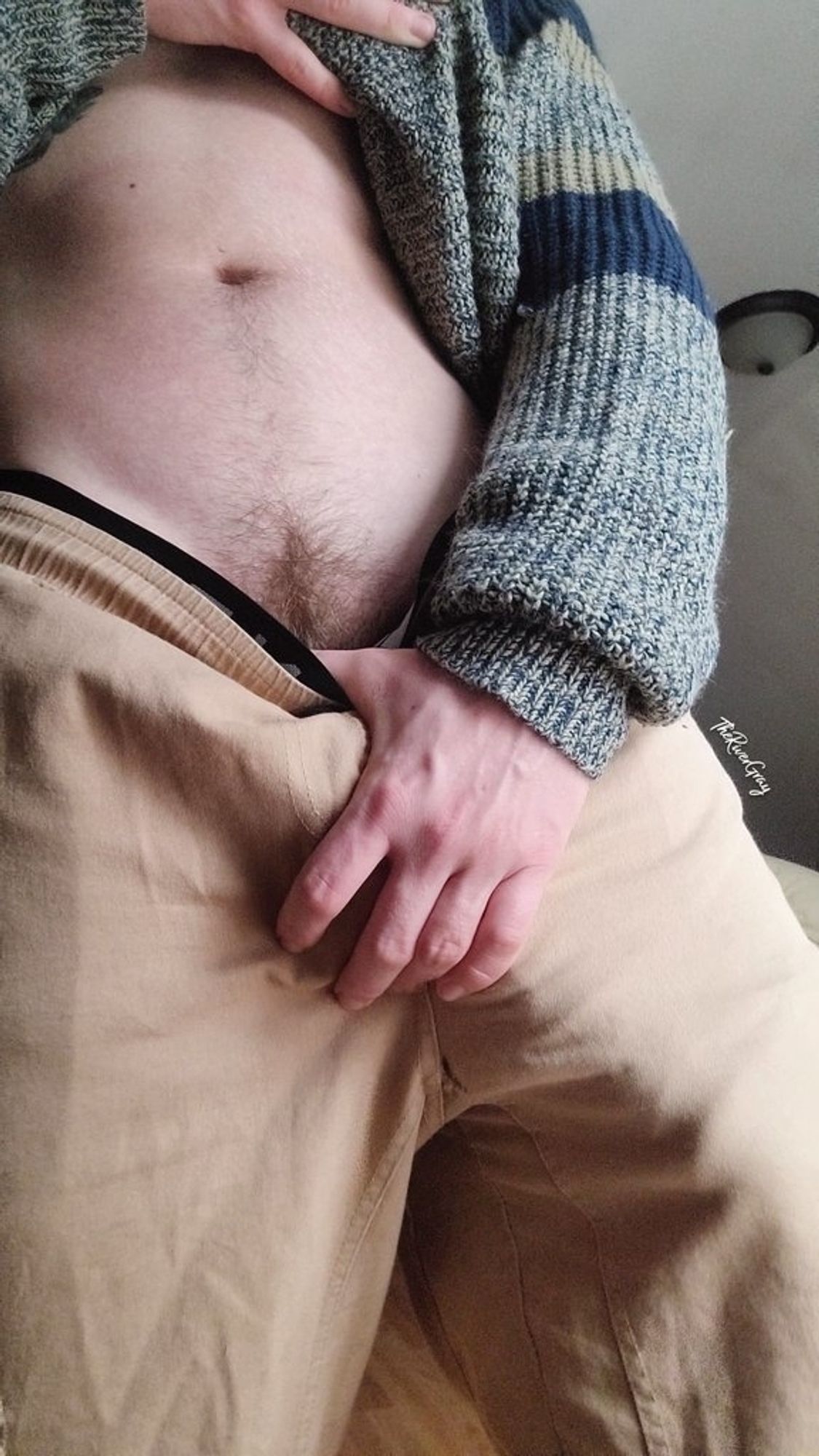 River lifting up his cozy sweater and pulling down his pants slightly to show off his happy trail and bush. The bulge of his hard cock showing through the tan pants.