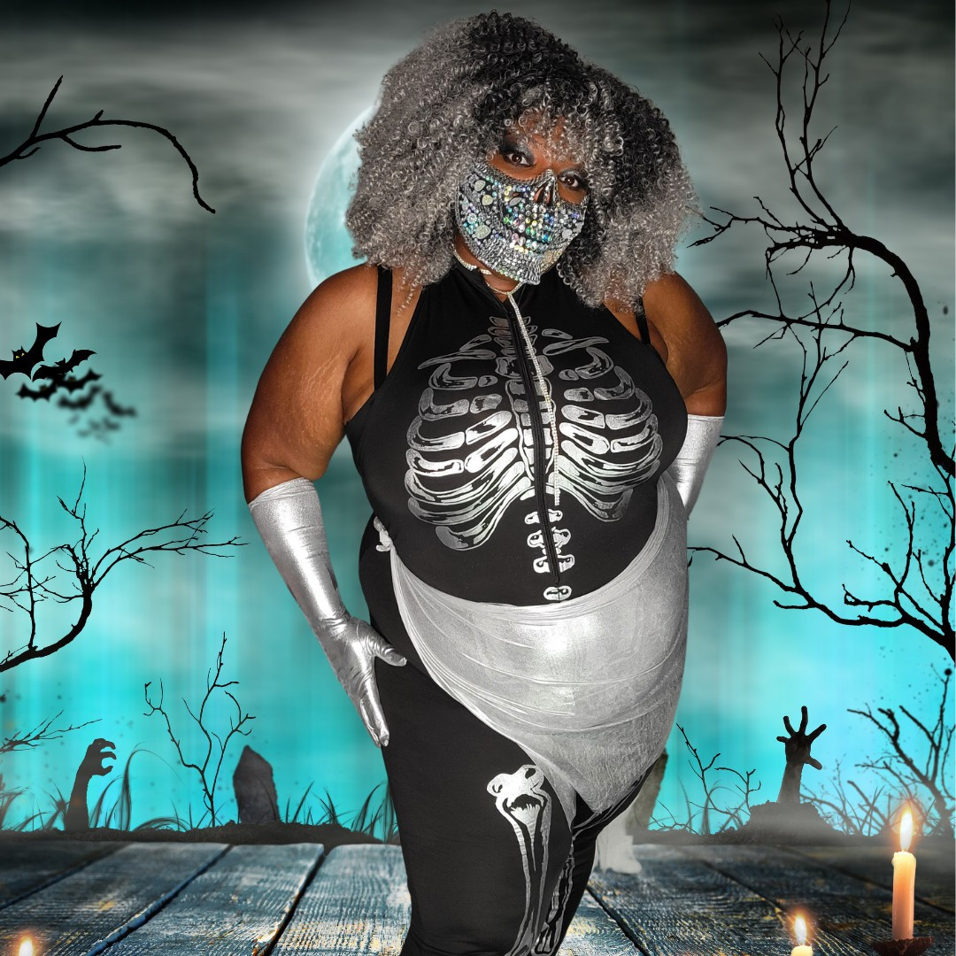 TaLynn Kel in a skeleton onesie wearing a bejewled skull mask on the bottom half of face. She has burly ombre grey/black hair and is wearing silver gloves and a silver sash and is standing in front of a faux graveyard.