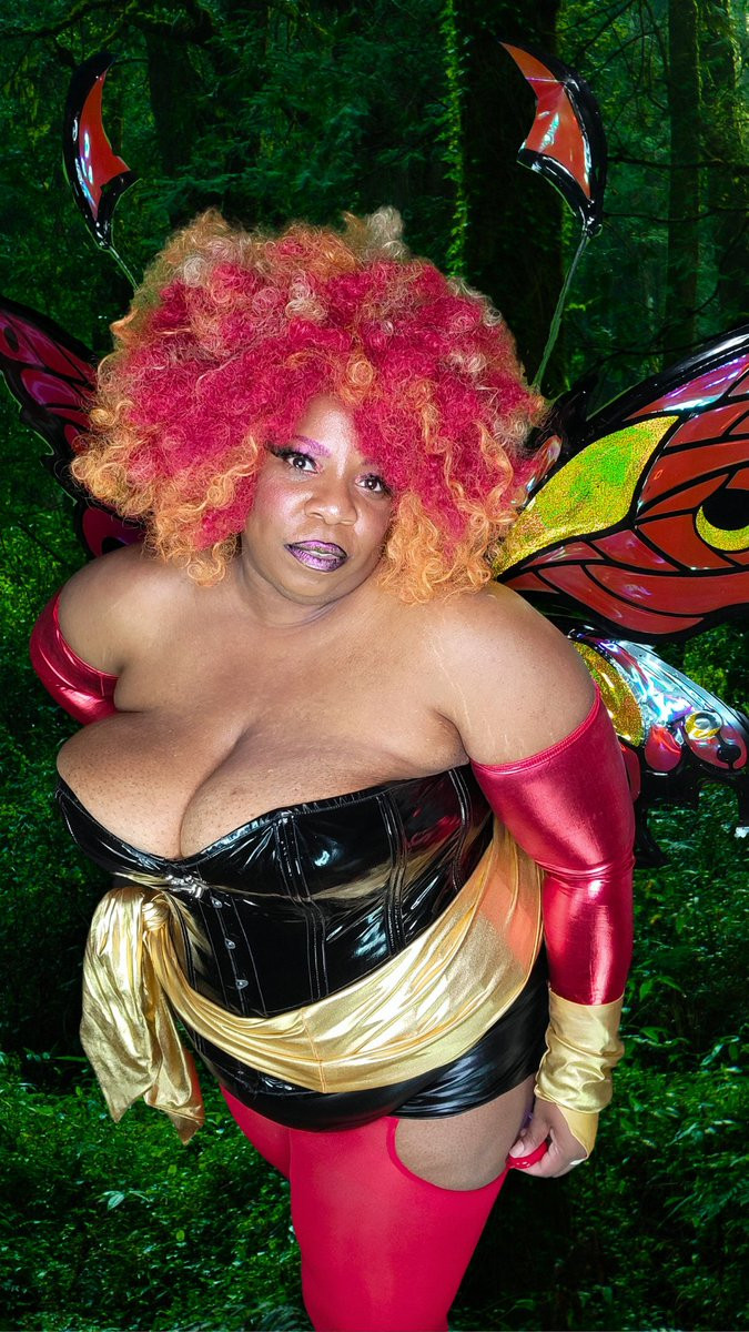 Full figured Black woman wearing a black vinyl corset, red sleeves, gold gloves, and a gold sash with a red and orange curly afro and red, black, and gold butterfly wings.