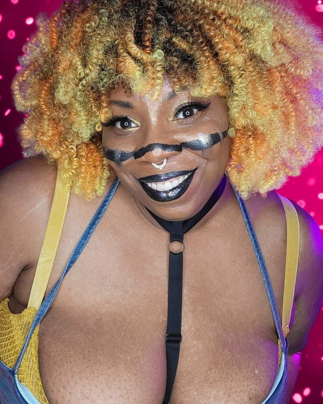 Image of a fat Black woman in a denim top and yellow bra, with orange, yellow and black curly hair. She has a black line painted across her face and black lipstick. She is smiling and looking at the camera.