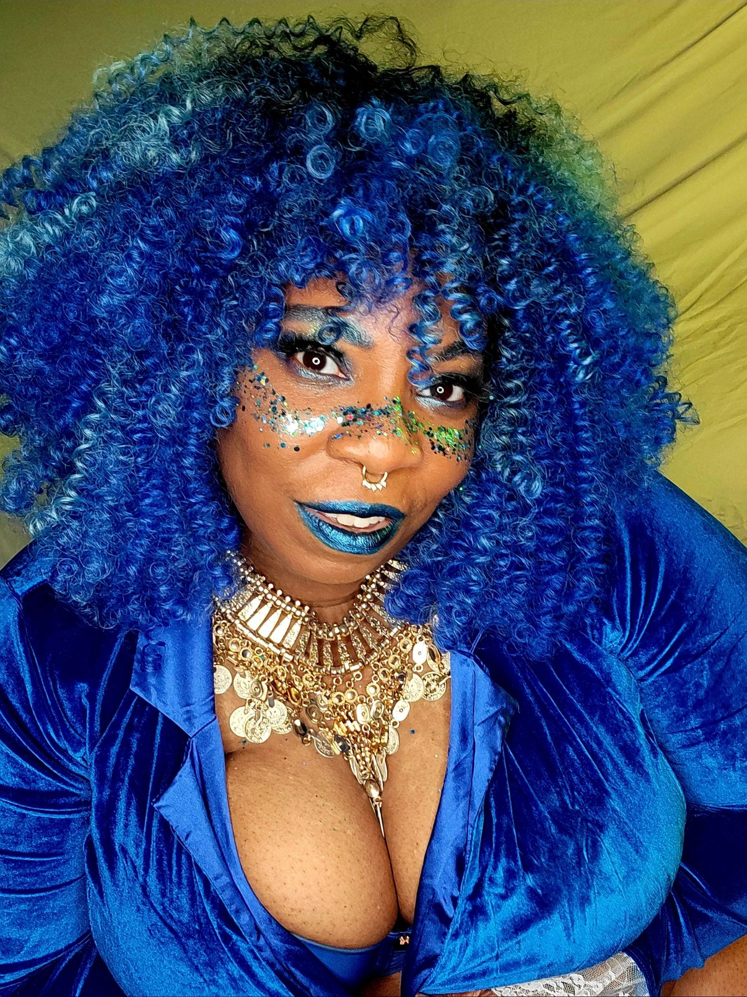 TaLynn Kel, a fat, Black woman, with blue curly hair, wearing blue makeup has glitter across her cherkbones and nose, is smiling at yhe csmera. She is wearing a blue velvet jacket and a chunky hold necklace.