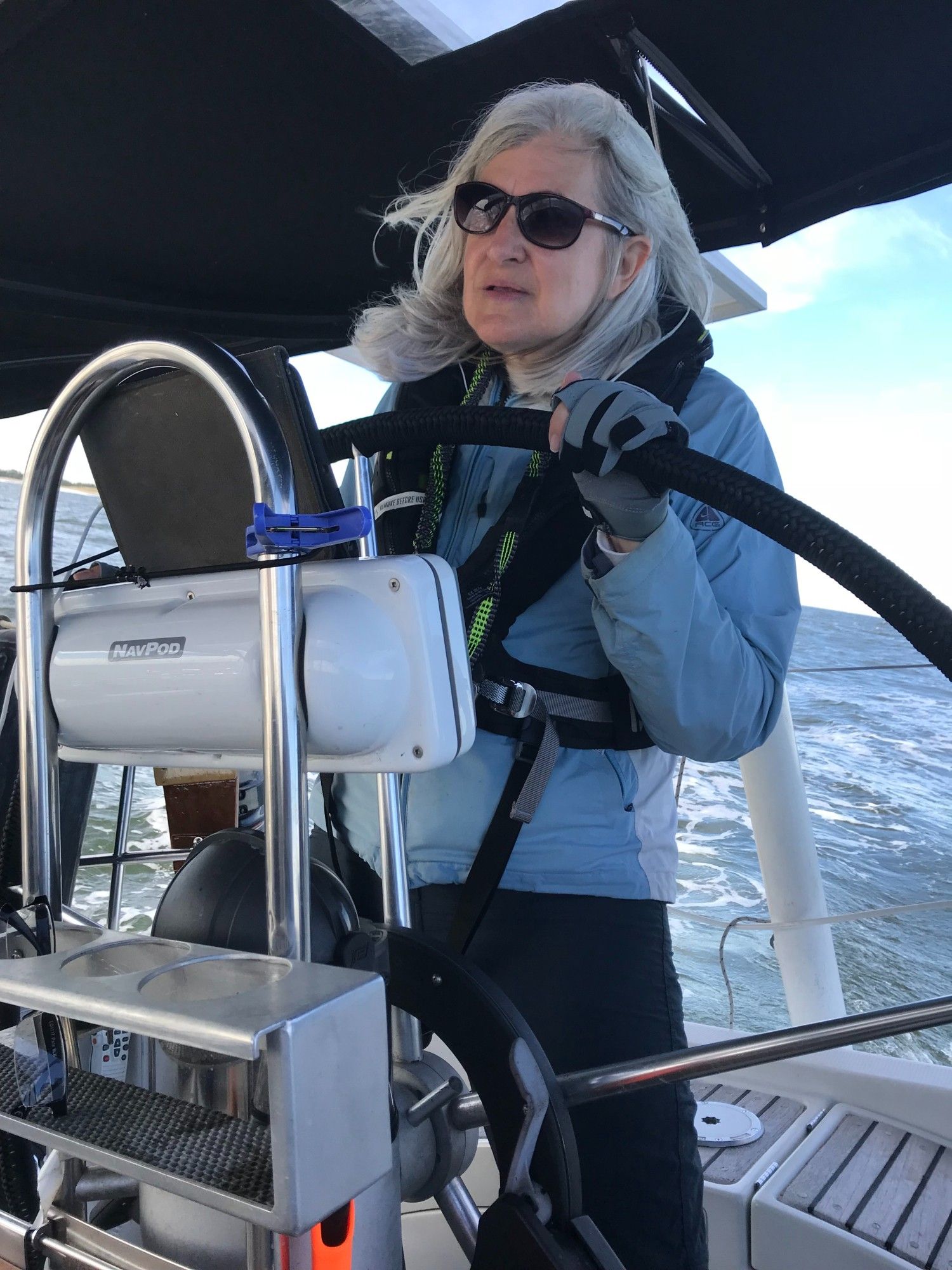 Picture of older woman with gray hair and jacket and life jacket on, body of water behind, at helm of sailboat.