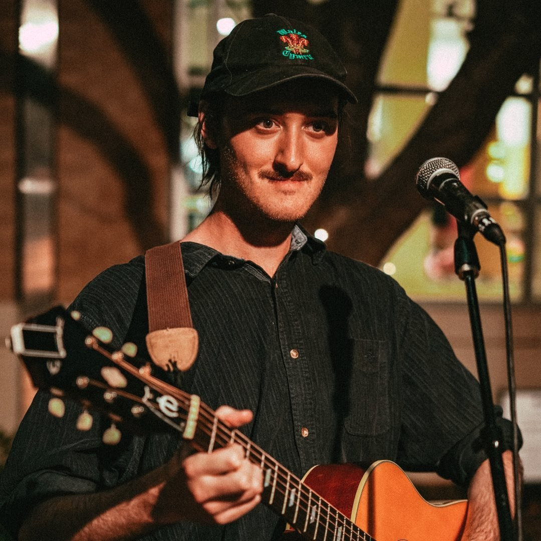 A close-up picture of me standing outside a community centre building in Texas the evening. I am smiling as I play an acoustic guitar with a microphone to the right of the picture. I am wearing a black Wales baseball cap, and a dark navy corduroy shirt.