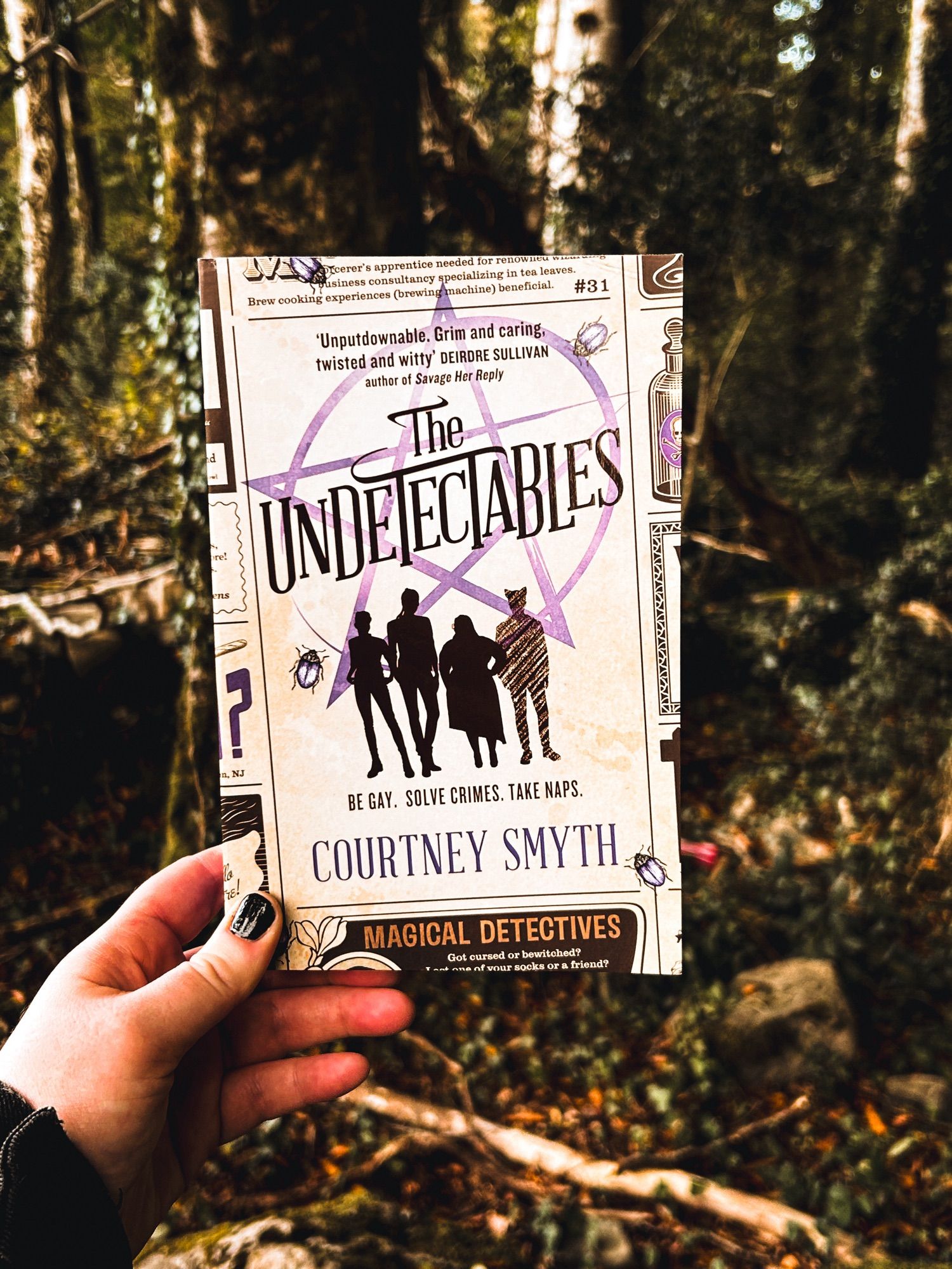 A white hand with black nail polish holds a copy of The Undetectables up against a forest scene, with autumnal trees, ivy and leaves in the background