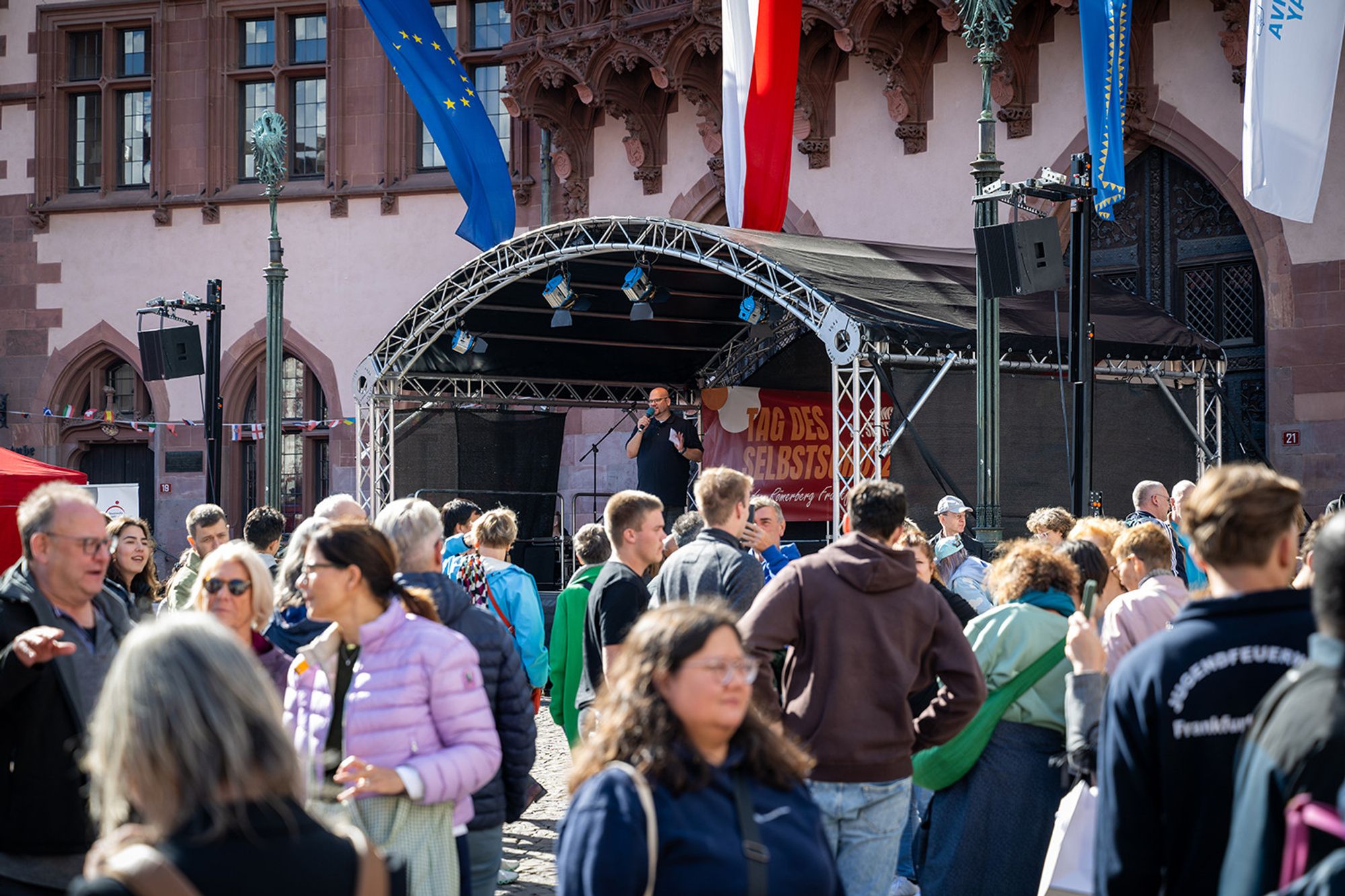 Der Blick schweift vom Römerberg über eine größere Menschenansammlung in Richtung einer Bühne, auf der Daniel R. Schmidt in ein Mikrofon spricht. Über der Bühne schweben die Fahnen des Römers, Frankfurts Rathaus.