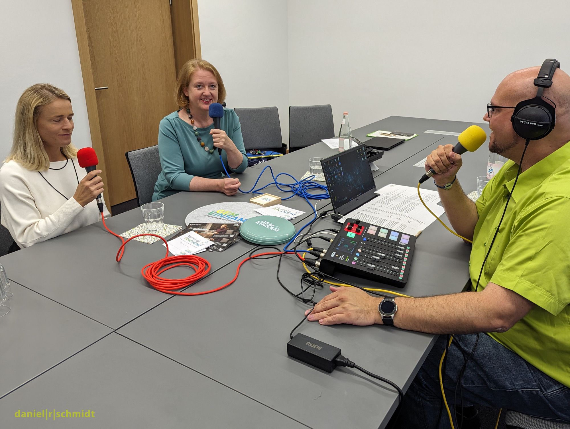 An sechs zusammengeschobenen Einzeltischen sitzen auf der linken Seite Verena Bentele und Bundesministerin Lisa Paus. Sie sprechen jeweils in Mikrofone. Auf der anderen Seite sitzt Podcasthost Daniel R. Schmidt und spricht ebenfalls in ein Mikrofon. Er sitzt vor einem Mischpult, einem Laptop-Computer und einem Redeskript.