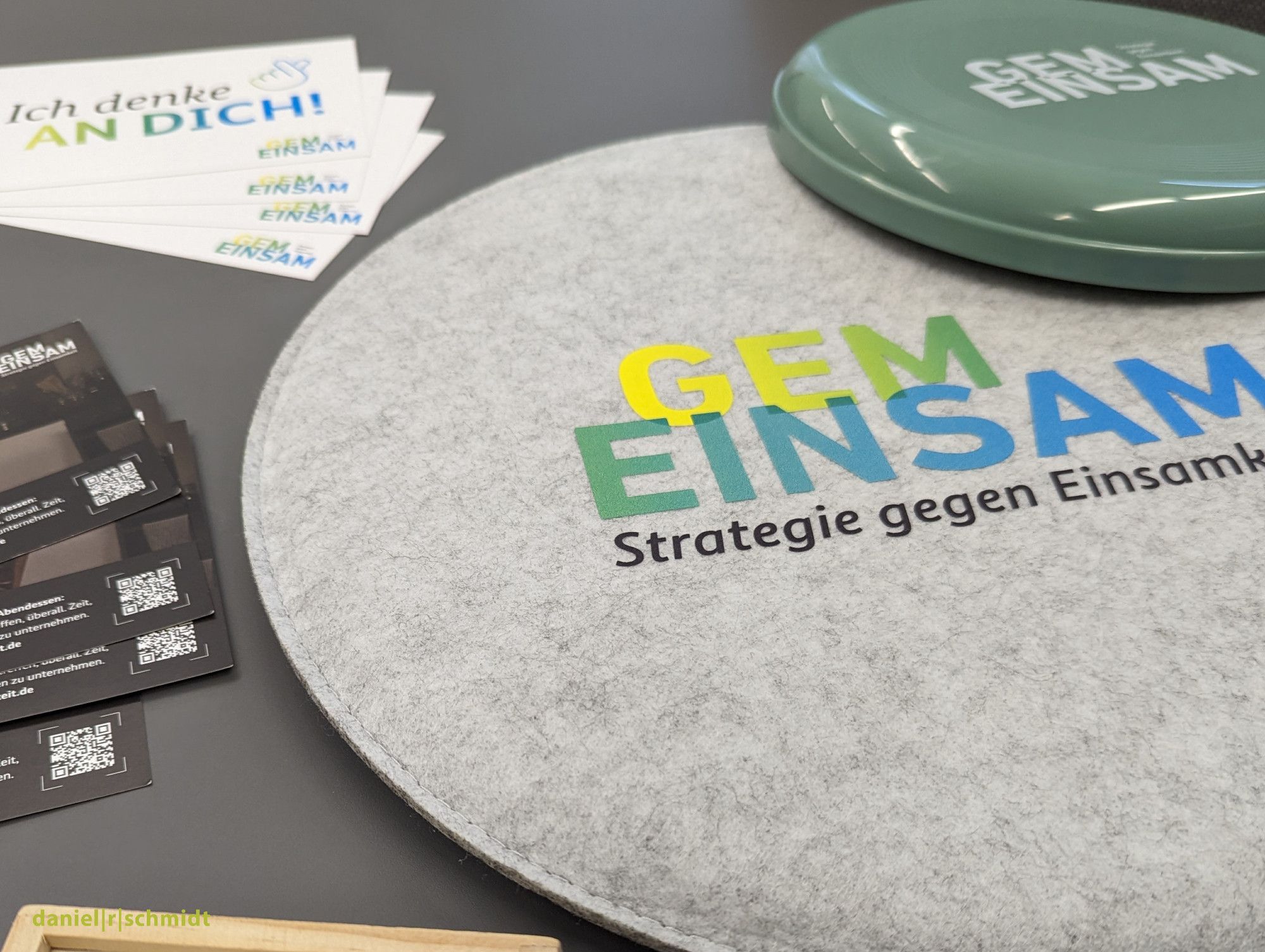 Auf einem Tisch liegen Werbematerialien der Bundeskampagne "Gemeinsam - Strategie gegen Einsamkeit". So unter anderem eine graue Spielscheibe, eine grüne Frisbee und weiße Aufkleber mit dem Schriftzug "Ich denke an Dich".