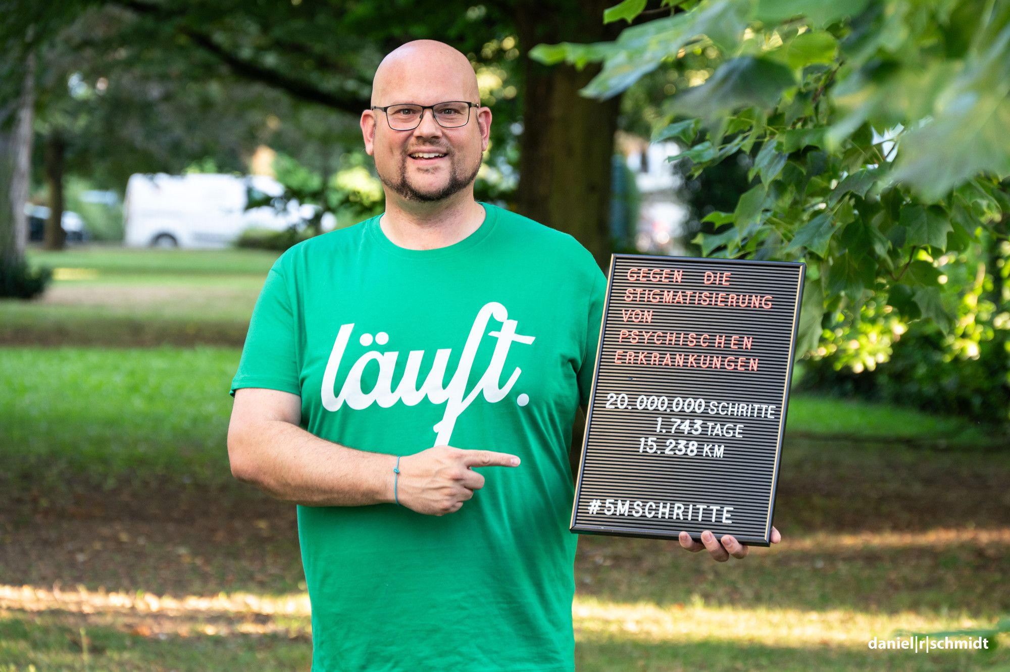 Daniel R. Schmidt steht in einem grünen T-Shirt mit der Aufschrift "läuft." vor Bäumen und grün, eingerahmt von parkenden Autos im unscharfen Hintergrund. Er hält eine schwarze Tafel in der Hand, auf die er zeigt. Die Tafel zeigt: "Gegen die Stigmatisierung von psychischen Erkrankungen. 20.000.000 Schritte, 1.743 Tage, 15.238 Kilometer".