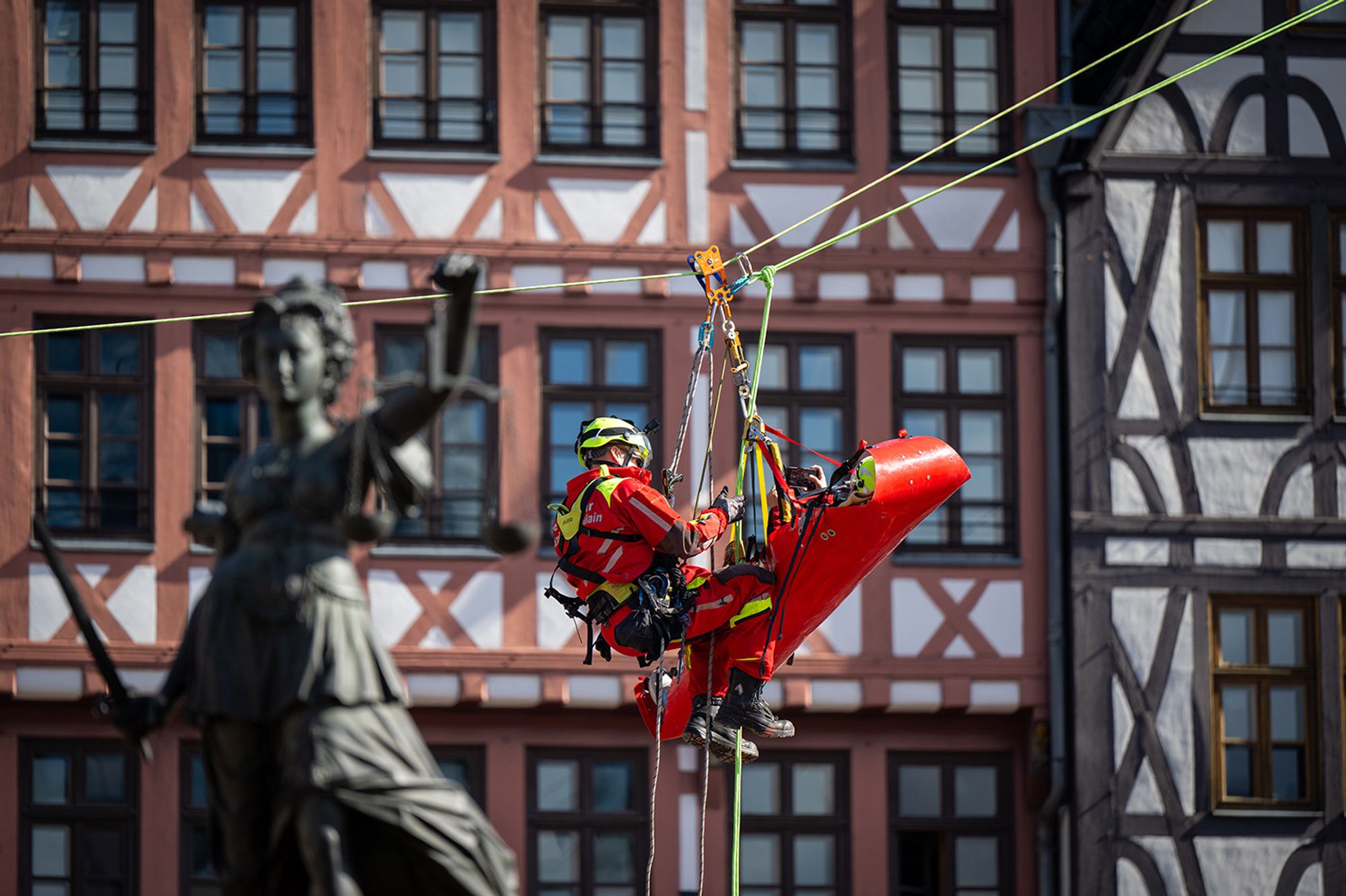 Ein Höhenretter der Feuerwehr Frankfurt seilt sich über den Römerberg mit einer Trage und Patientin darin ab. Im Vordergrund ist unscharf die Justitia des Gerechtigkeitsbrunnens zu erkennen.