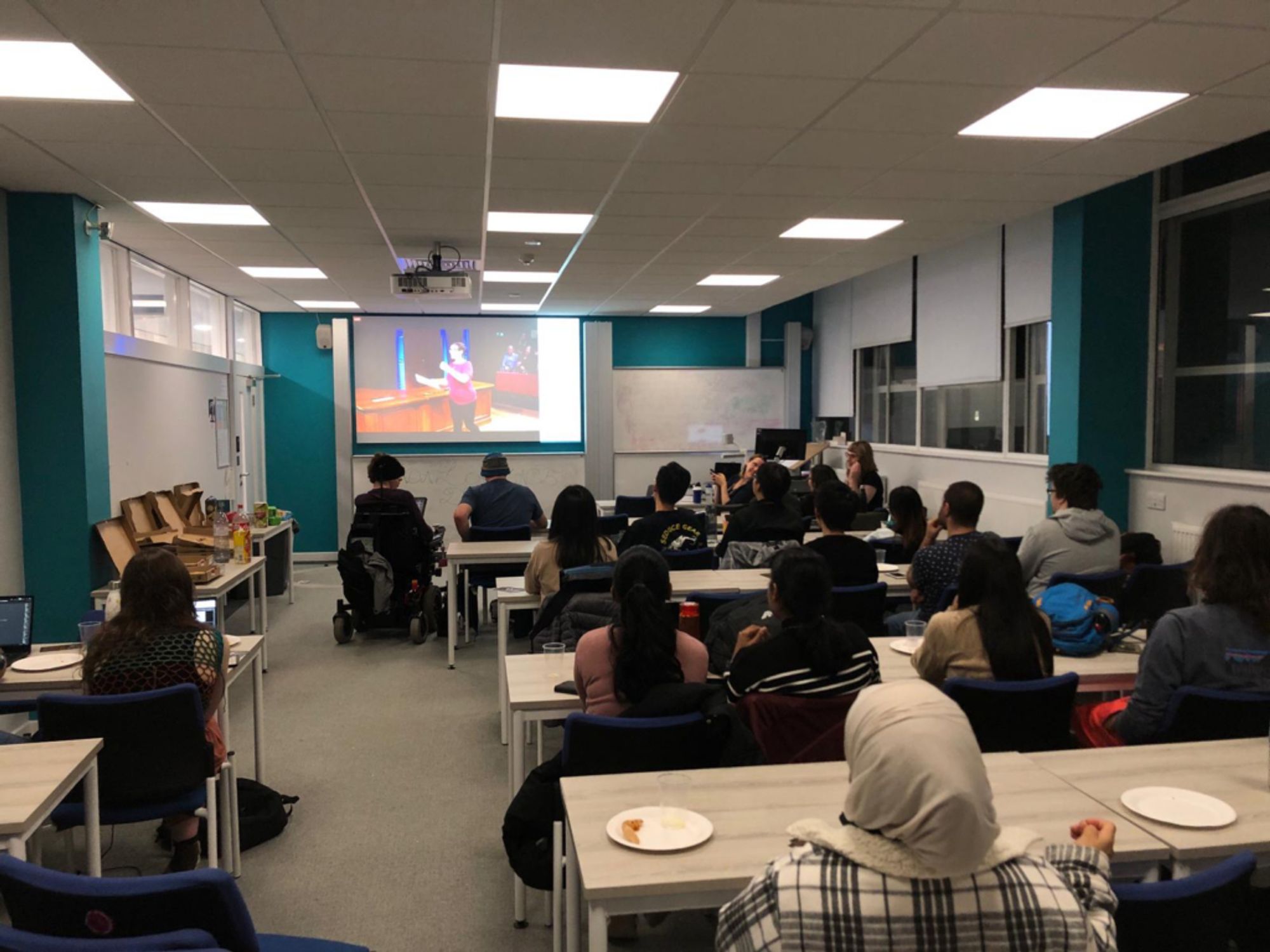 Students and staff watching a livestream of the Royal Institution’s Ada Lovelace Day Live ‘science cabaret’, celebrating the contributions of women in science.

The attendees are sitting in rows next to many pizza boxes, facing away from the camera and towards the livestream on a projector screen.