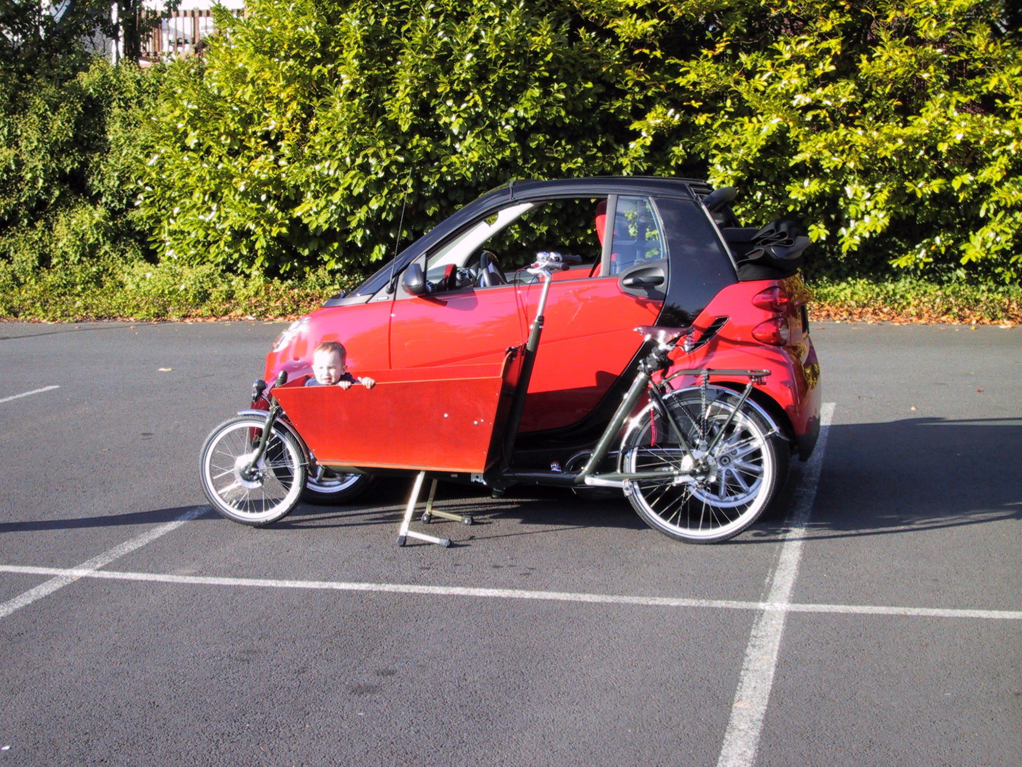 A red smart fortwo with black tridion panel and roof in a parking lot. In front of the smart is a cargo bike that's nearly as long as the car. Sitting in the wooden front cargo carrier of the bike is a small child whose head pokes over the top.