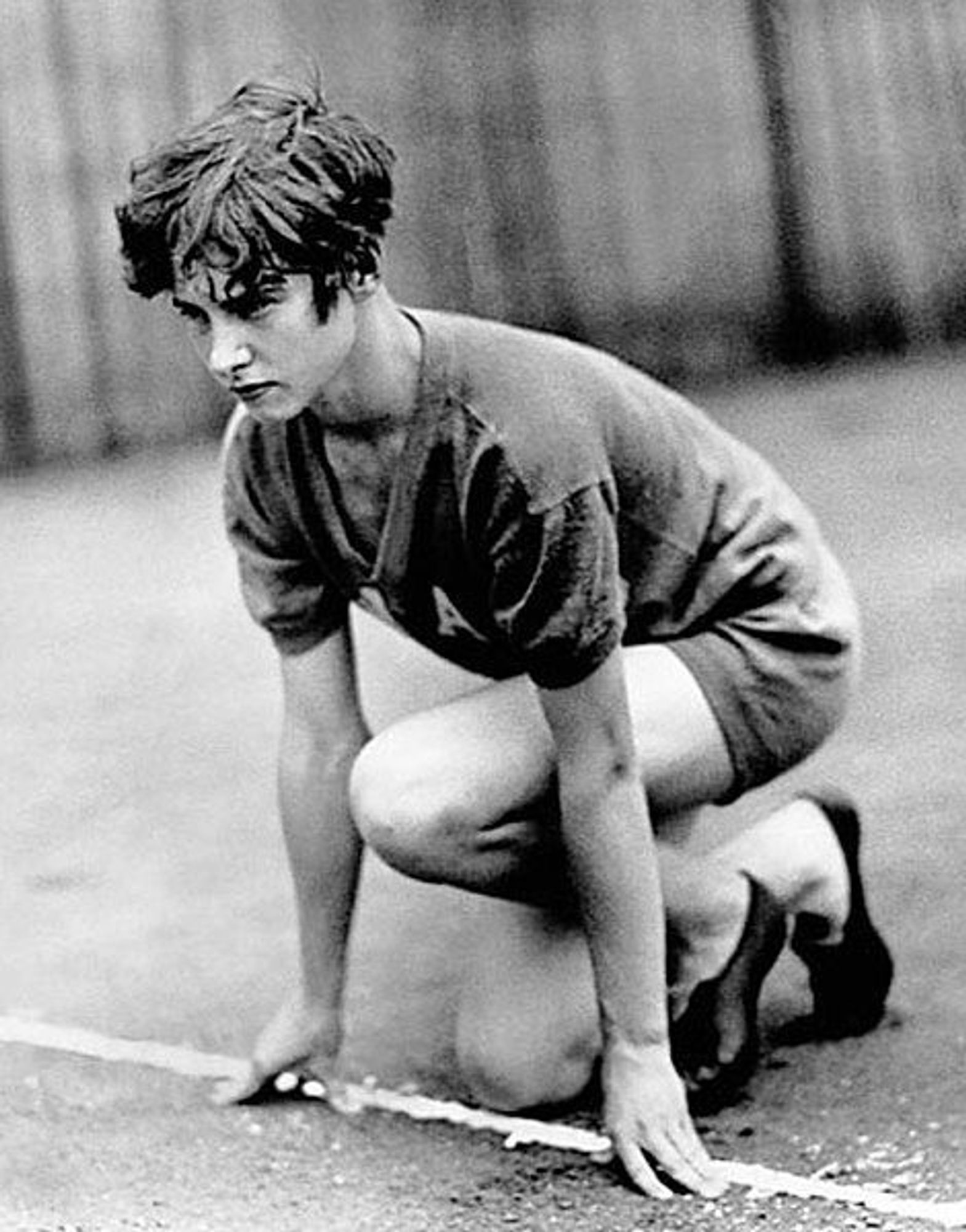 Photo en noir et blanc de Betty Robinson, cheveux courts, accroupie sur une piste, prête à commencer une course. Elle porte un t-shirt et un short, et ses mains sont posées sur le sol devant elle.