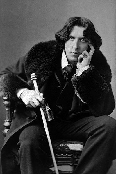 Photo en noir et blanc d'Oscar Wilde. Portrait de trois quarts, de face, assis, penché en avant, le coude gauche posé sur le genou, la main au menton, tenant une canne dans la main droite, portant un manteau.