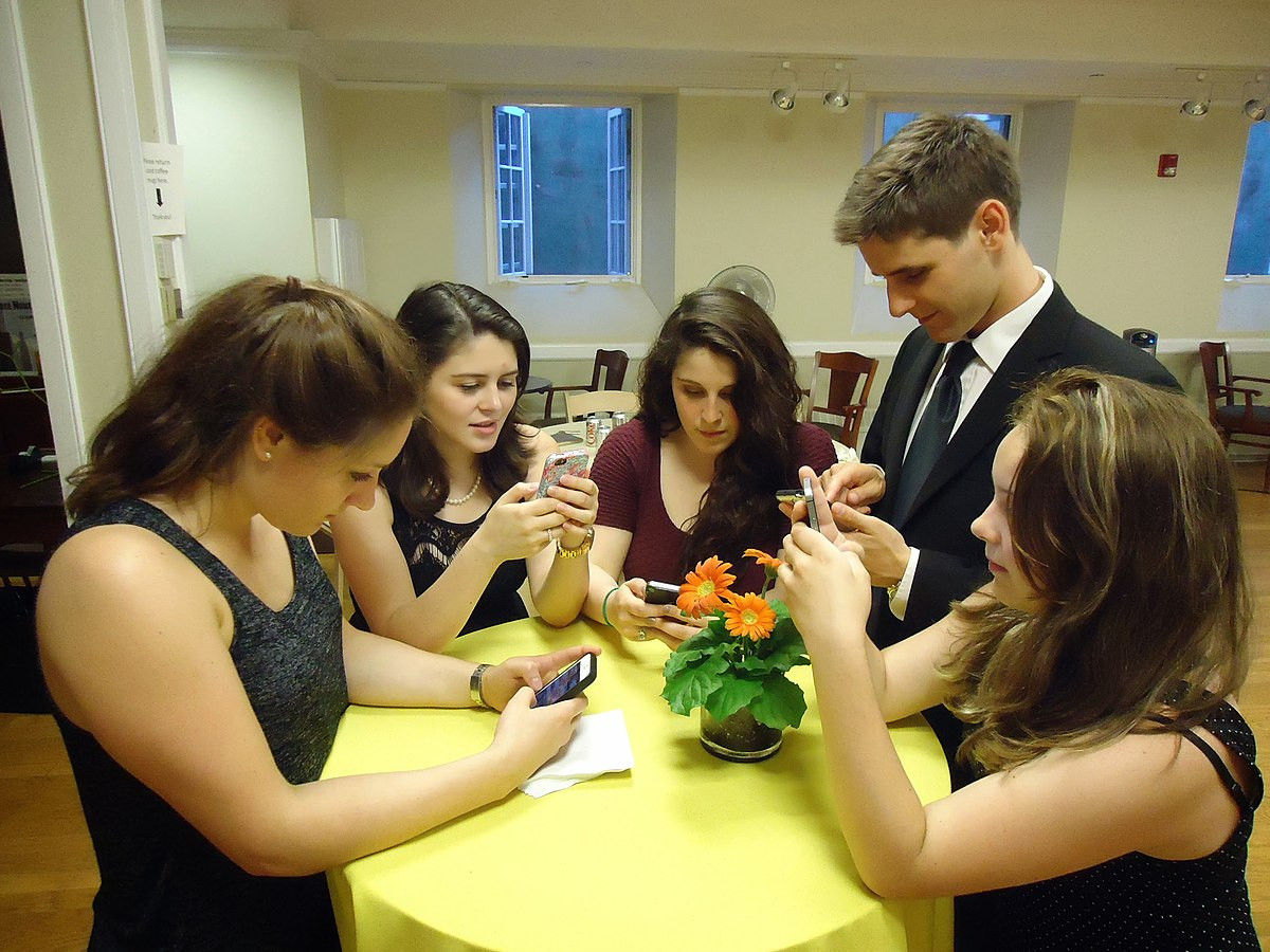 Photo d’un groupe de cinq jeunes (4 filles et un garçon) lors d'une fête, debout autour d’une table jaune avec un petit pot de fleurs orange au centre. Chacun est concentré sur son smartphone, suggérant une scène sociale moderne où la technologie est omniprésente.
