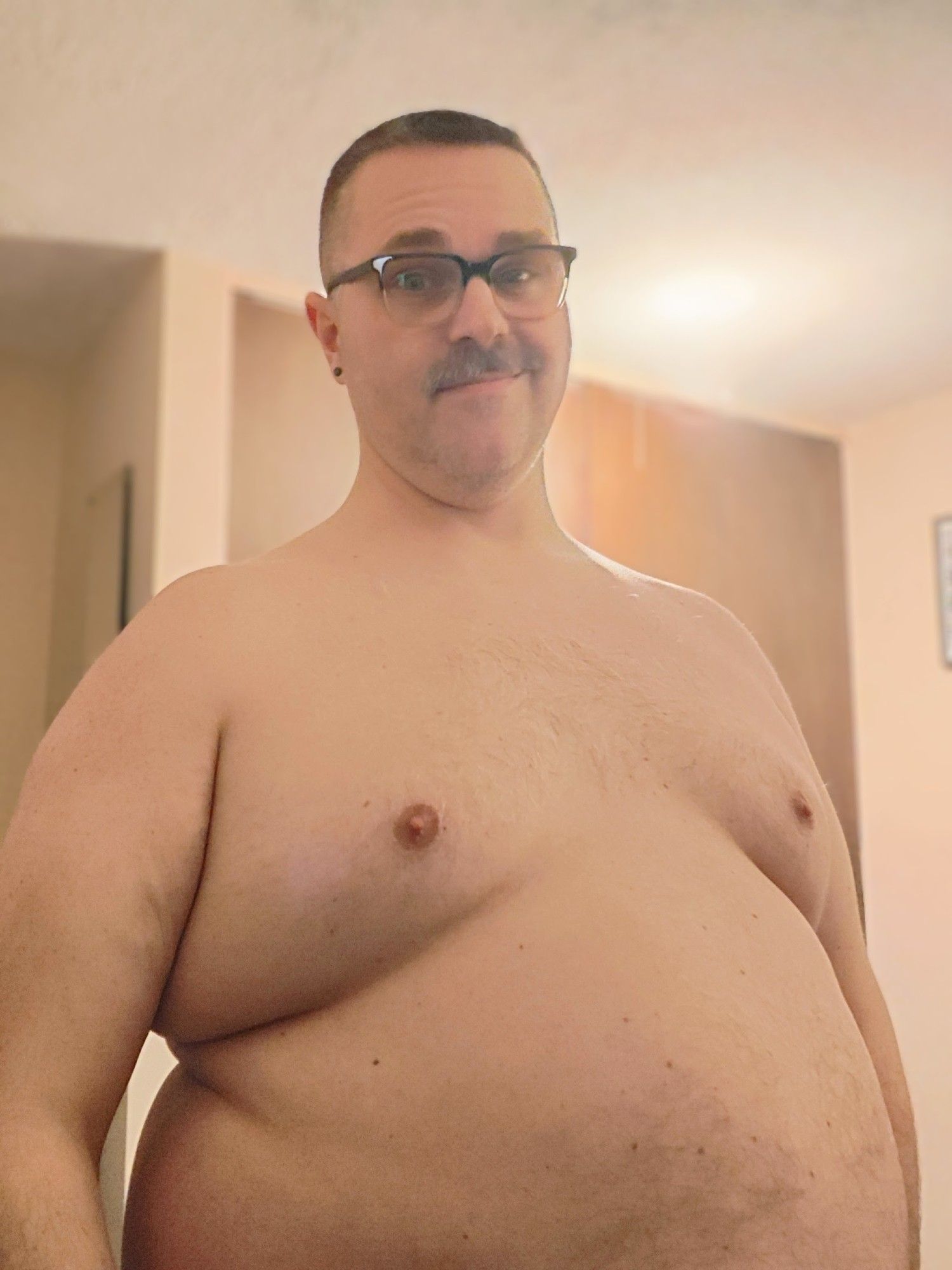 A shirtless fat man standing by himself. He's wearing black framed glasses and is smirking, ish.