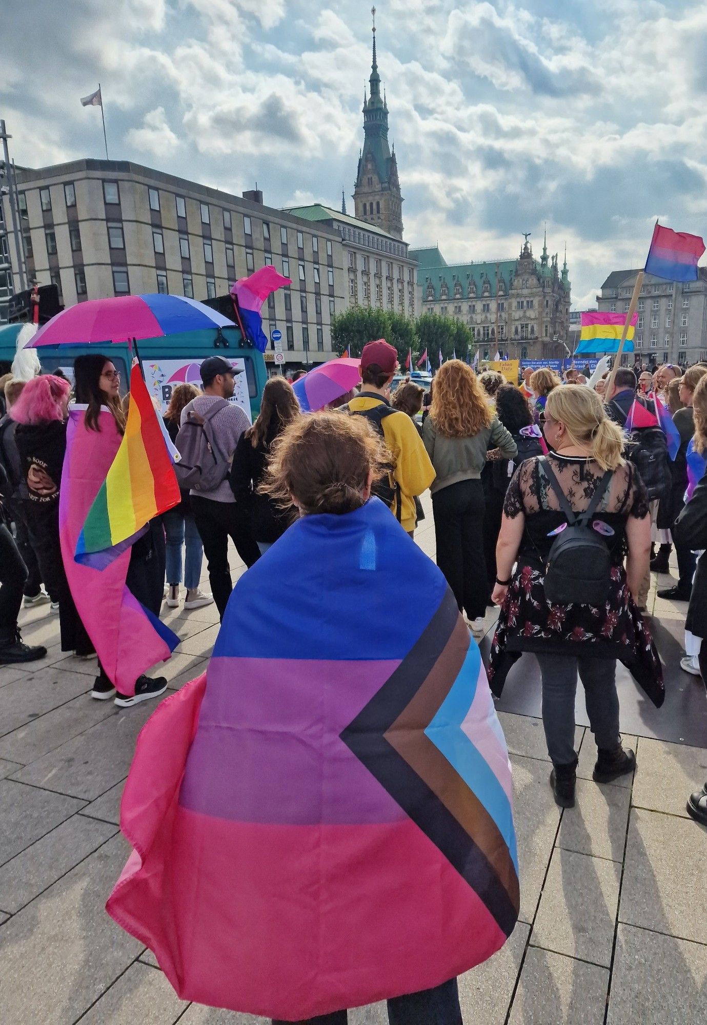 Crowd listening to a speech at the bi+ pride march today. Picture is shot from the back, lot's of bi, pan and rainbow flags visible. I'm seen from behind, wrapped in a progressive bi flag