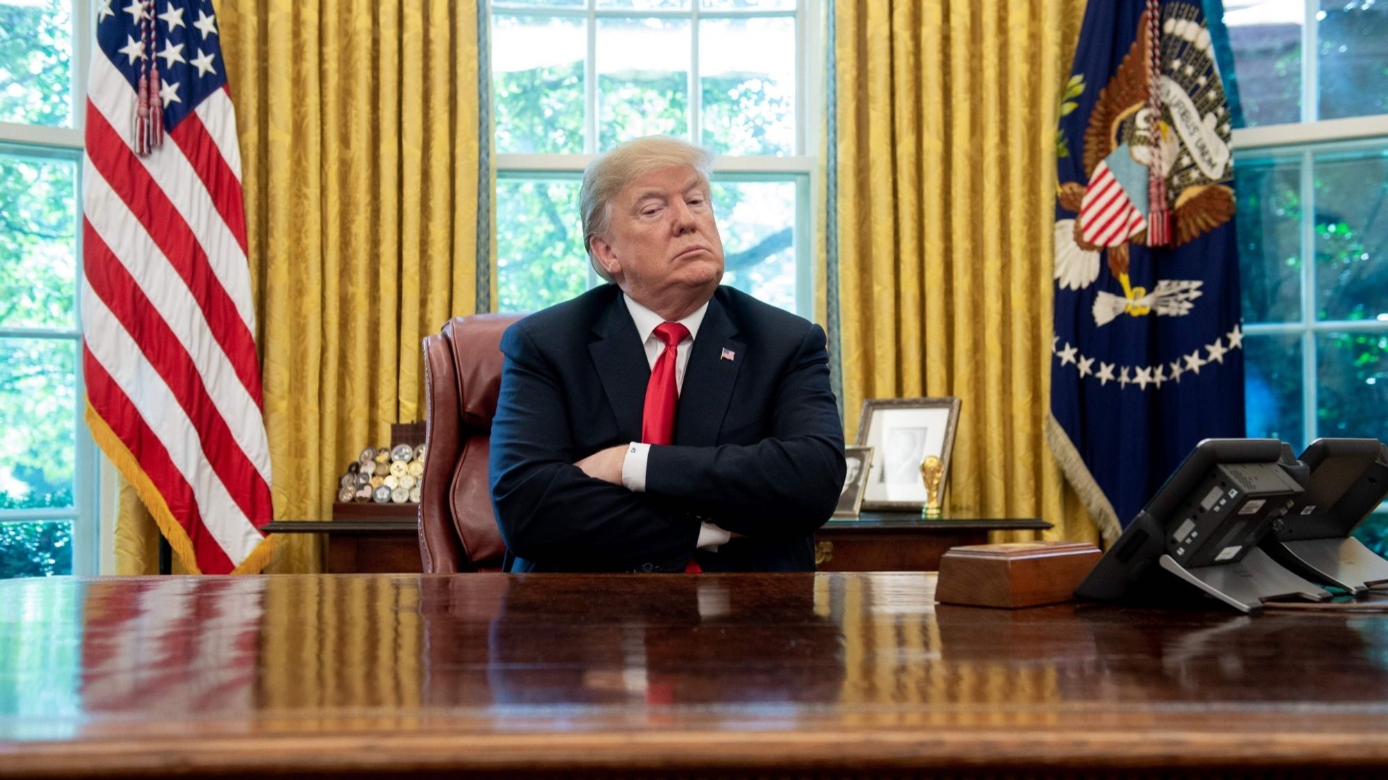 Foto Querformat 
Donald Trump Sitz im Ovaloffice hinter einem Schreibtisch verschränkt die Arme und schaut trotzig drein. Hinter ihm drei Fenster mit einer gelben offen Gardinen. Links und rechts ist Trump eingerahmt von amerikanischen Flaggen.