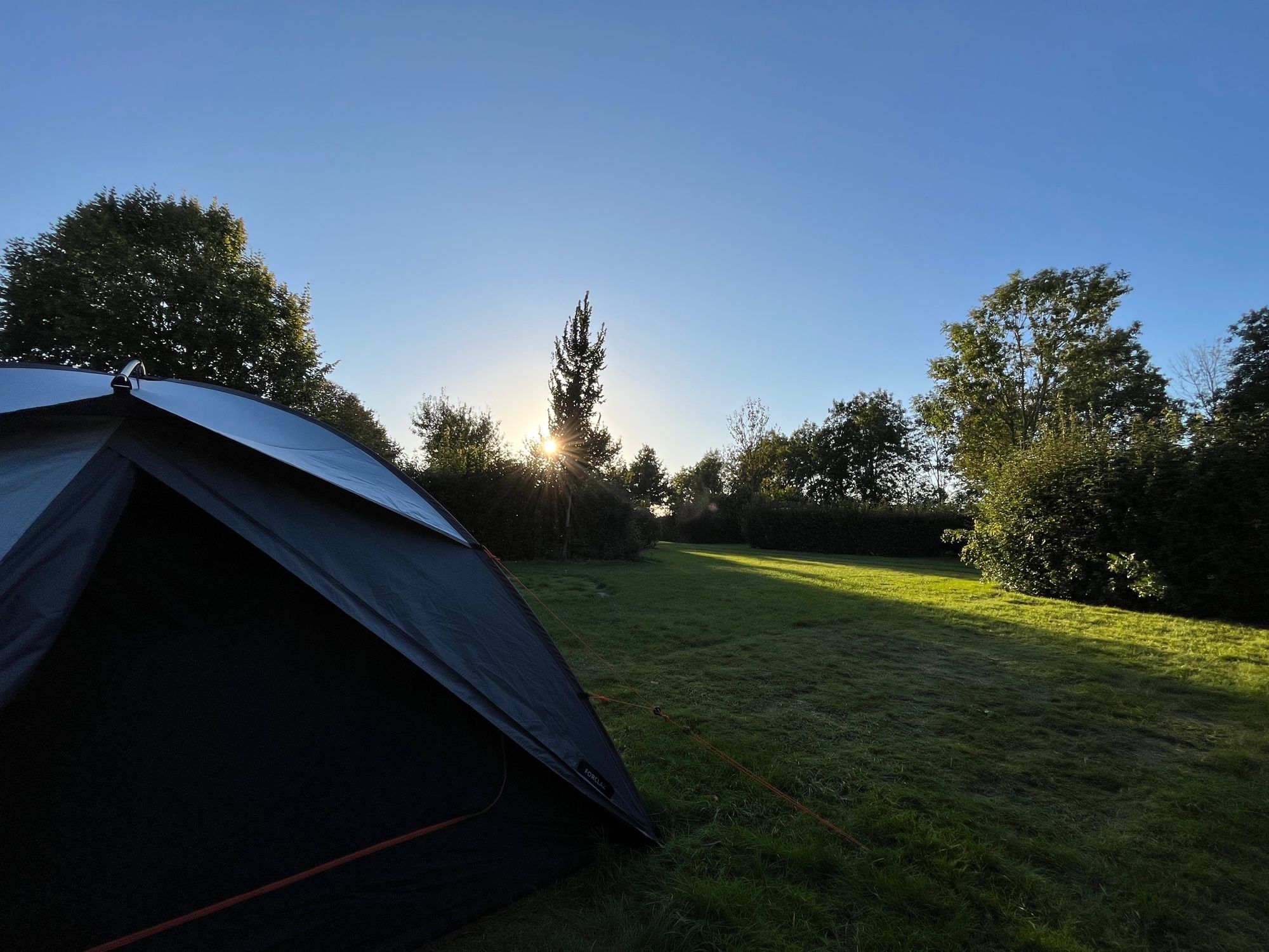 Kleine tent op grasveld en bomen met ondergaande zon. 