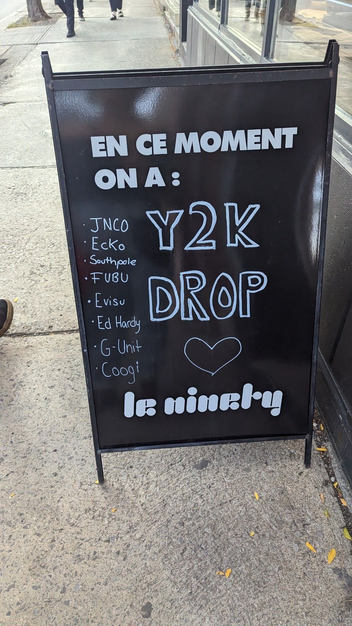 A black sandwich board sign with writing that says En Ce Moment on a: 

Y2K drop

JNCO, Ecko, Southpole, FUBU, Evisu, Ed Hardy, G-Unit and Coogie 

Le Ninety