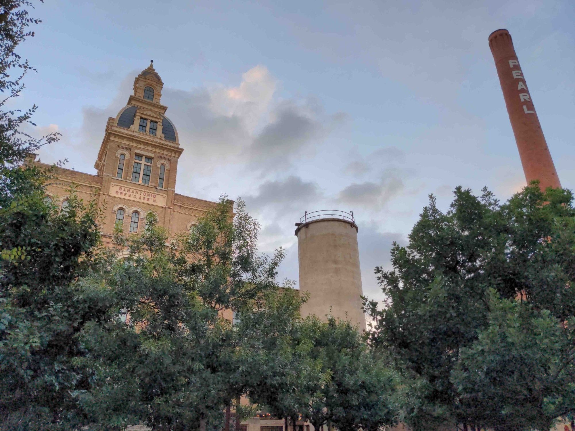 Trees in the foreground. Background has historic buildings, structures, and smokestack of the once Pearl Brewery against a partly cloudy sky.