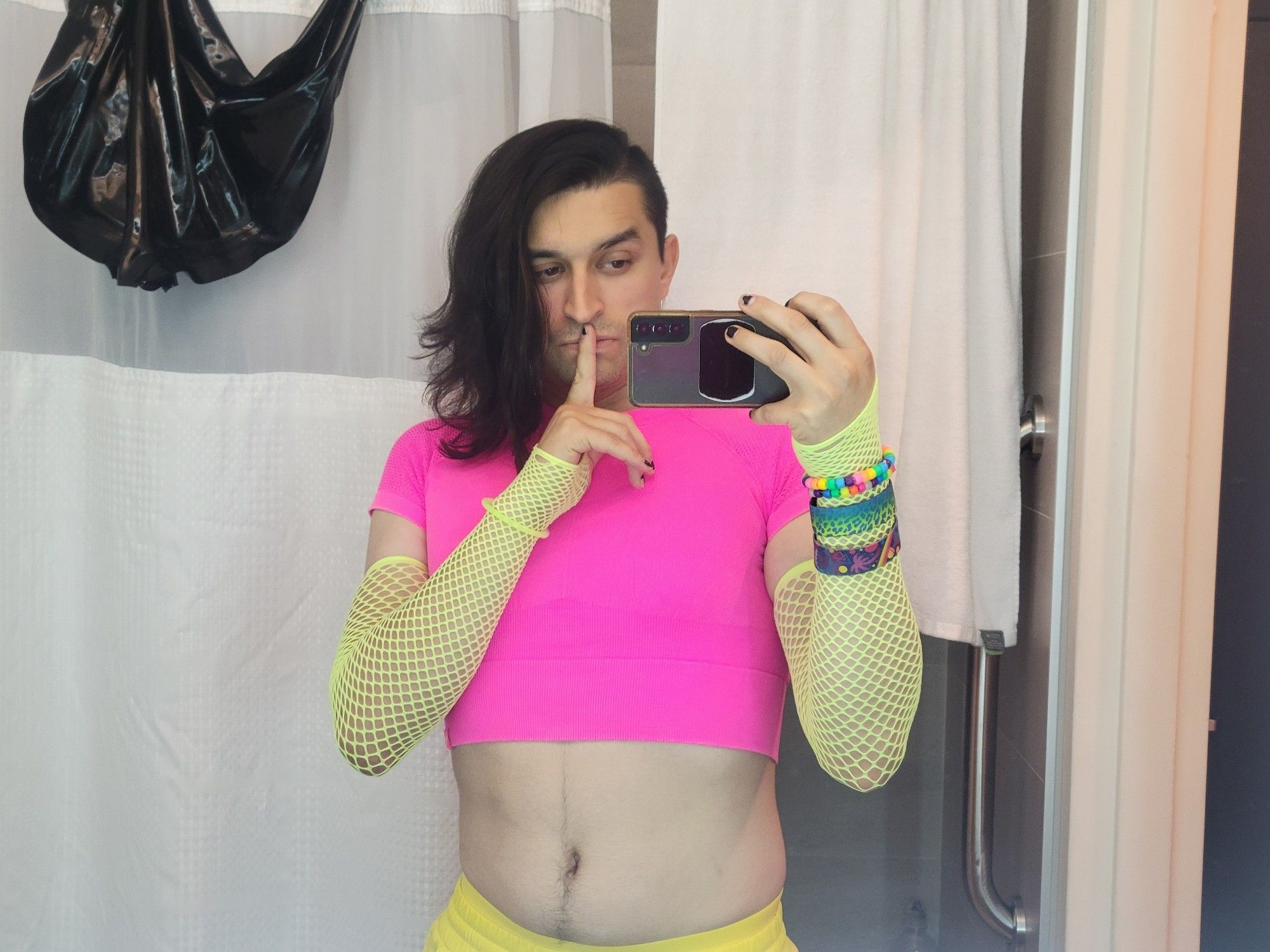 A hotel bathroom selfie of myself. My hair is down and clipped to the side, I'm wearing a yellow skort, hot pink crop top thing, neon green fishnet sleeves, and a few colorful bracelets. I have my finger in front of my lips in a "shh" pose, just because it looks good.