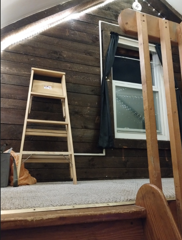 view of a loft room from the stairs. the camera is at floor level pointed up at the left side of a window on the dark brown far wall. white rope lights are hung providing illumination