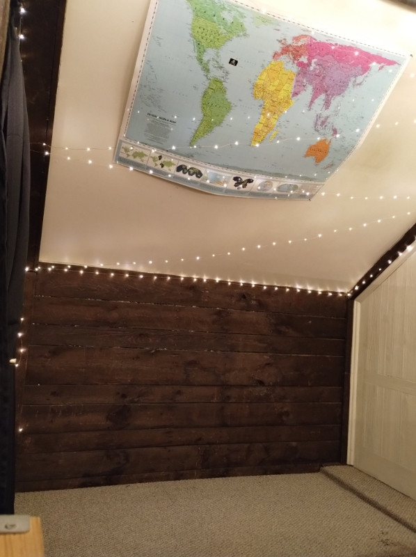 a low dark brown wall with a white ceiling over at a 45 degree angle. there's a white faux wood paneled wall to the right. a world map and fairy lights are hung on the ceiling
