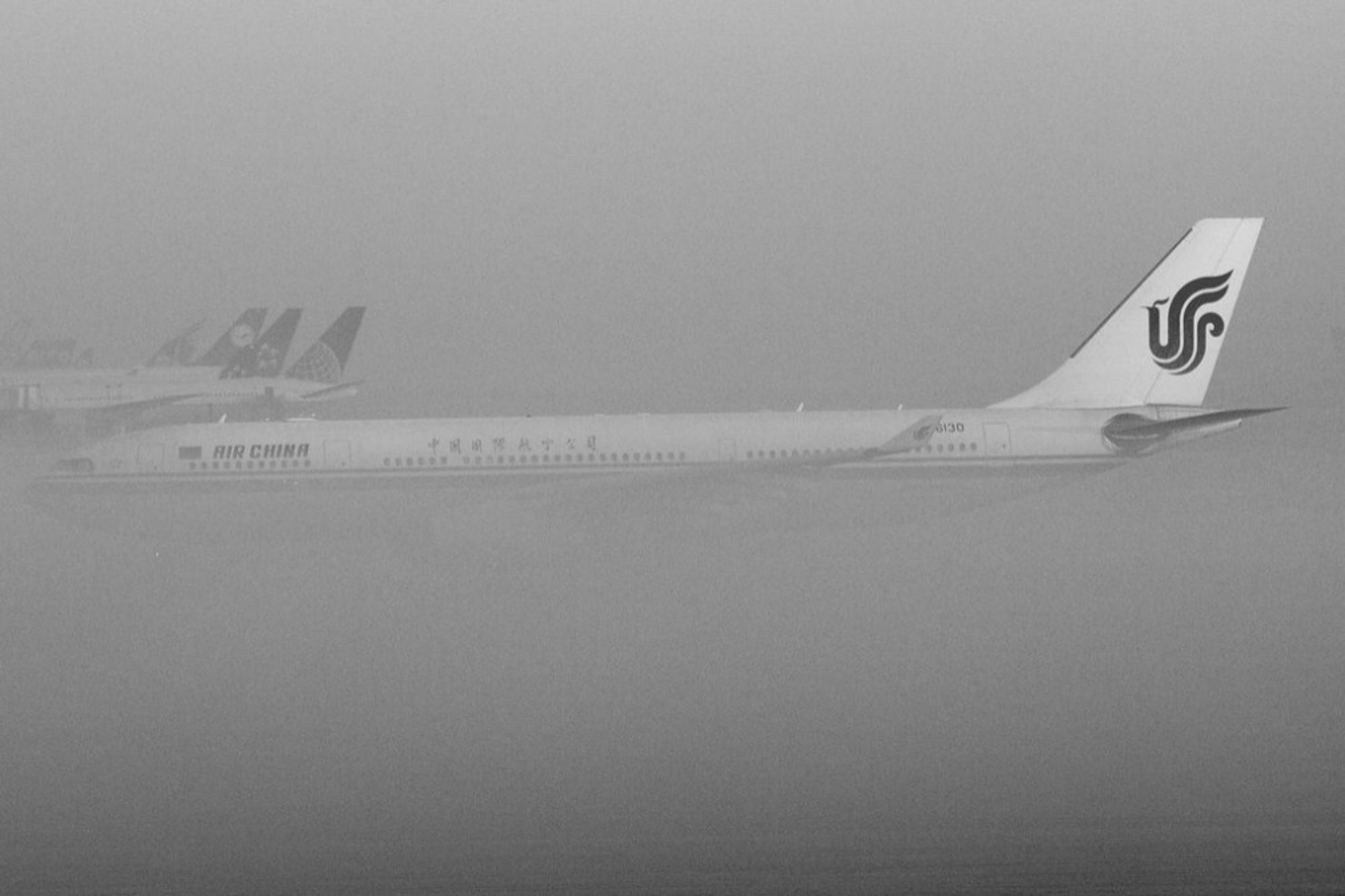 Das Foto zeigt mehrere Flugzeuge am Boden eines Flughafens, die von dichtem Nebel umgeben sind. Im Vordergrund ist ein Großteil eines Flugzeugs der Fluggesellschaft Air China zu sehen. Nur der hintere Teil des Flugzeugs ist deutlich erkennbar, einschließlich des  Logos der Fluggesellschaft. Die anderen Flugzeuge im Hintergrund sind durch den Nebel weniger detailliert und wirken eher wie Silhouetten.